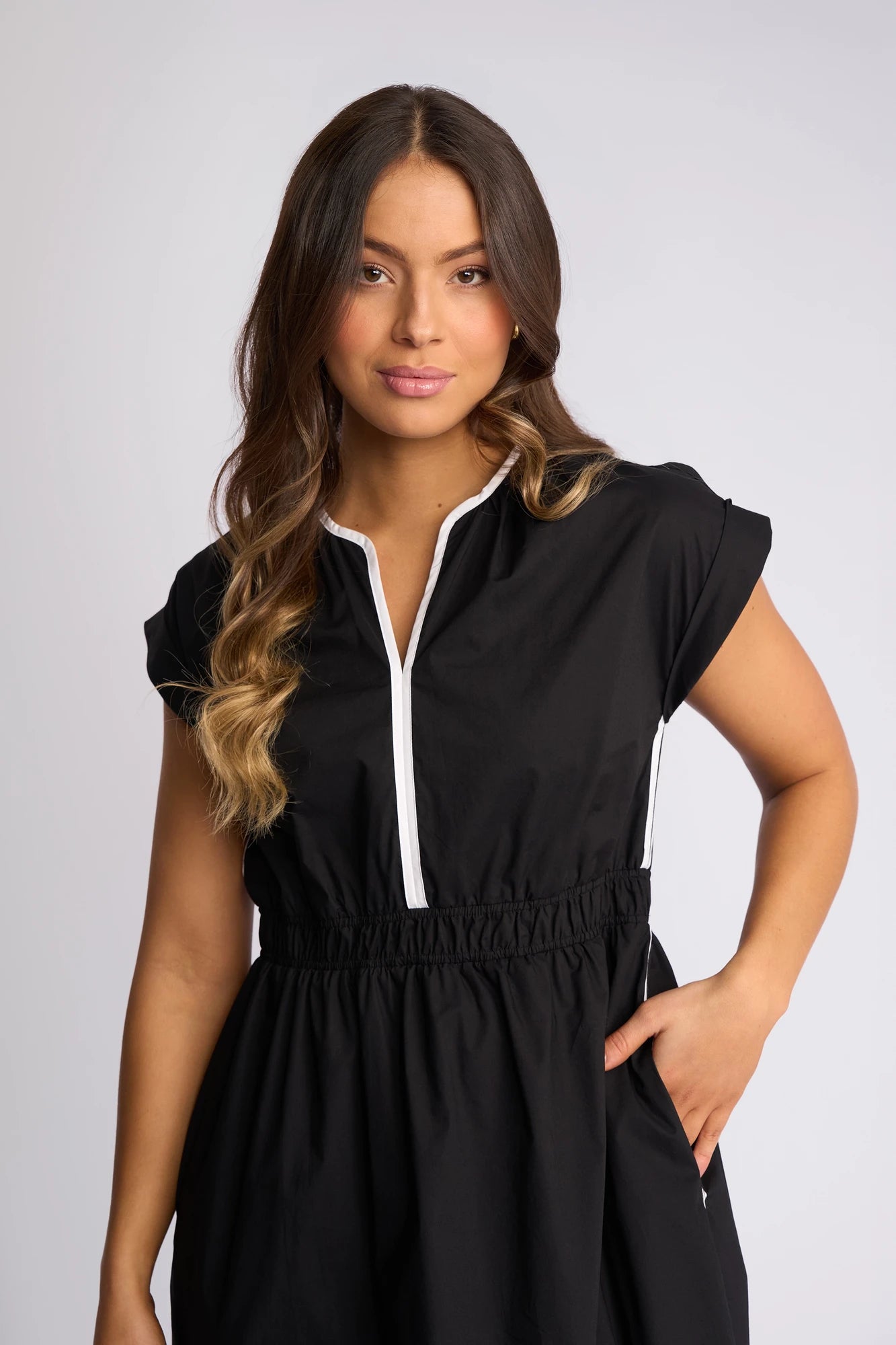 A woman with long, wavy brown hair wears the Vassalli Emmie Dress in Black/White, a relaxed-fit cotton midi dress with white trim. She stands against a plain light background, smiling softly and placing one hand in her pocket, effortlessly showcasing the breathable comfort and stylish ease of the Emmie Dress.