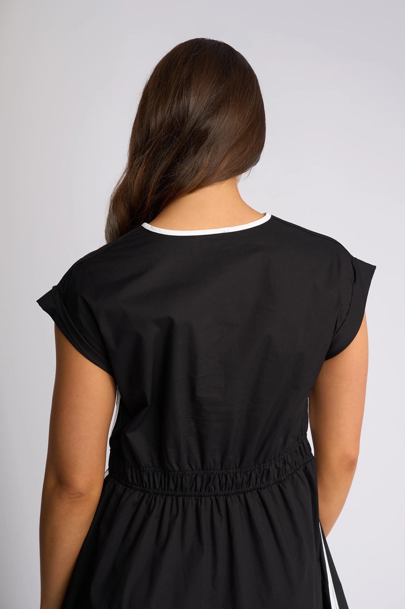 A person with long brown hair is shown from the back against a plain light background, wearing the Vassalli Emmie Dress in Black/White, which features white trim, a relaxed silhouette, and a midi length.