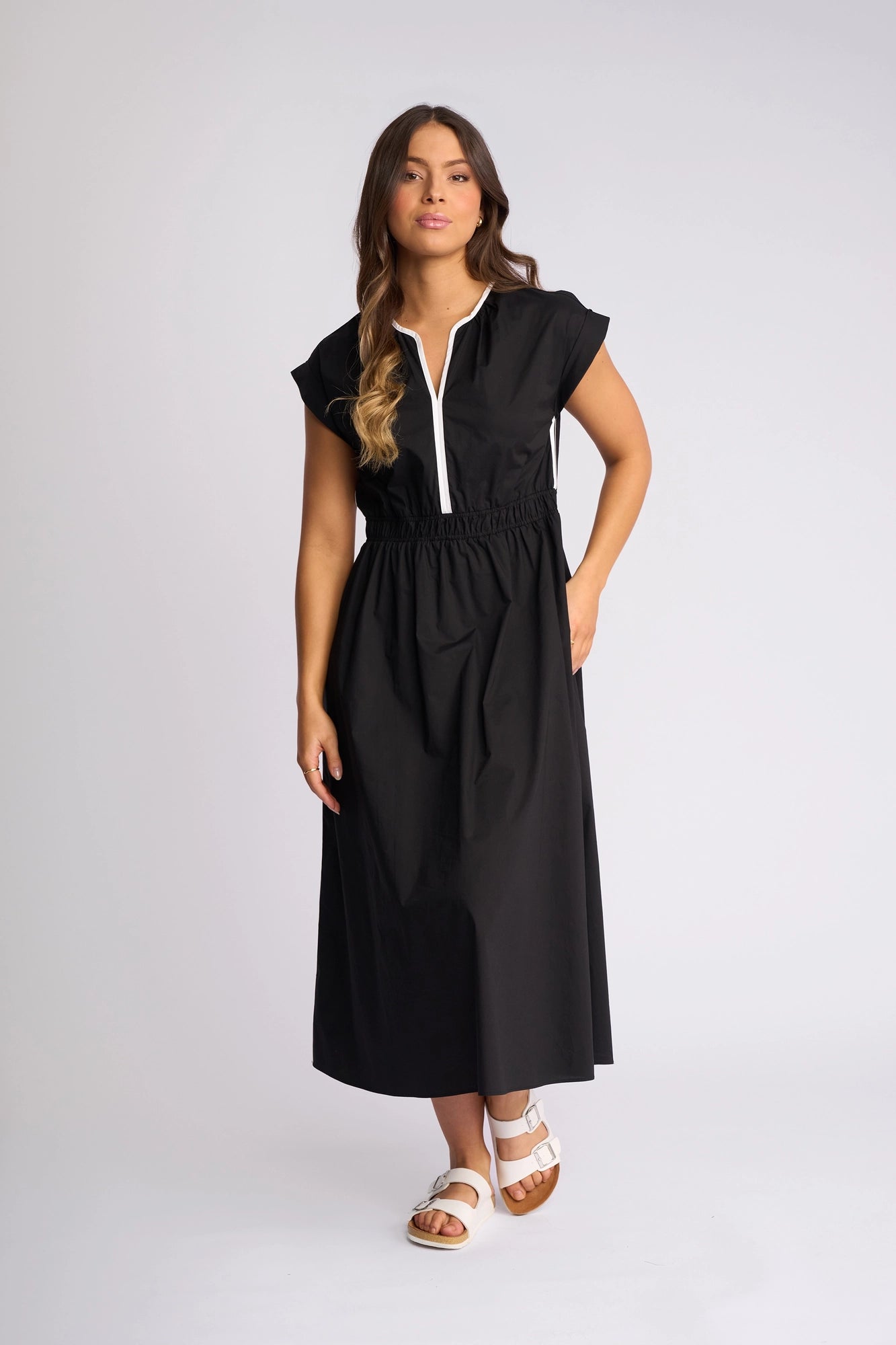 A woman stands against a plain background wearing the Vassalli Emmie Dress in Black/White—an ankle-length, short-sleeve cotton midi dress featuring a white zipper. The relaxed silhouette and breathable fabric pair well with her white sandals, long brown hair, and neutral expression.