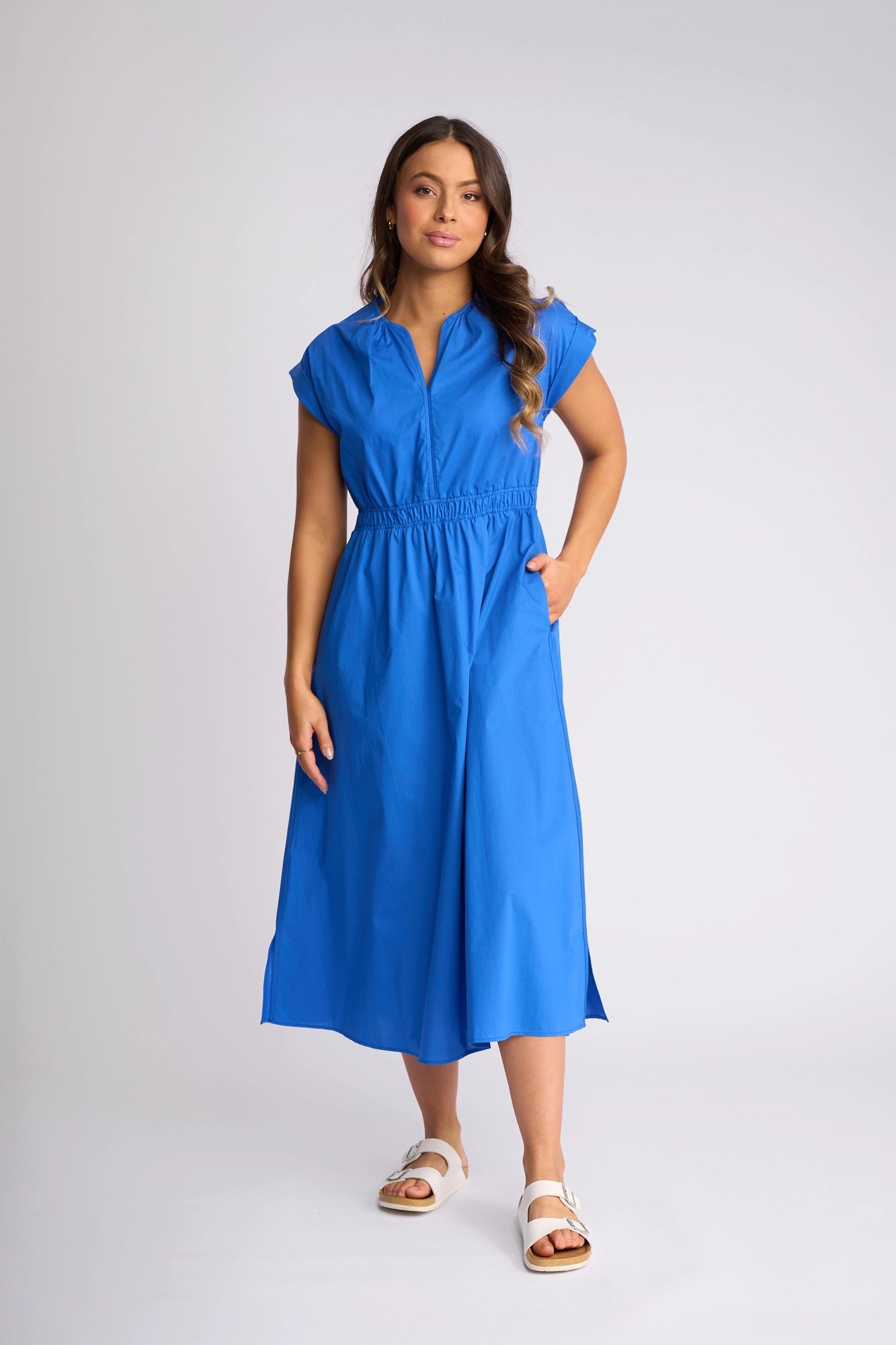 A woman stands against a plain background wearing the Vassalli Emmie Dress in cobalt—a bright blue, short-sleeve cotton midi dress featuring a gathered waist and relaxed, breathable silhouette. Styled with white sandals and one hand in her pocket, she wears her long hair in loose waves.