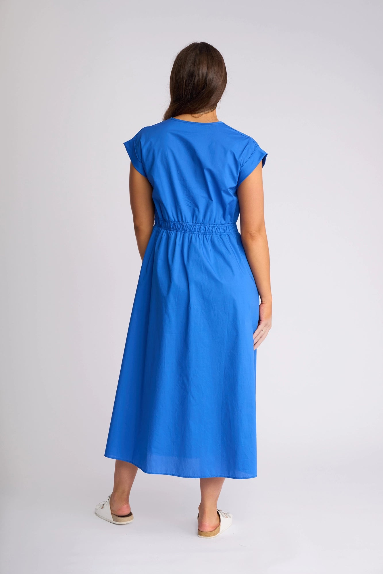 A woman stands facing away from the camera, wearing the Vassalli Emmie Dress in Cobalt—a bright blue cotton midi dress with a cinched waist and relaxed silhouette—paired with white flat shoes, all set against a plain light background.