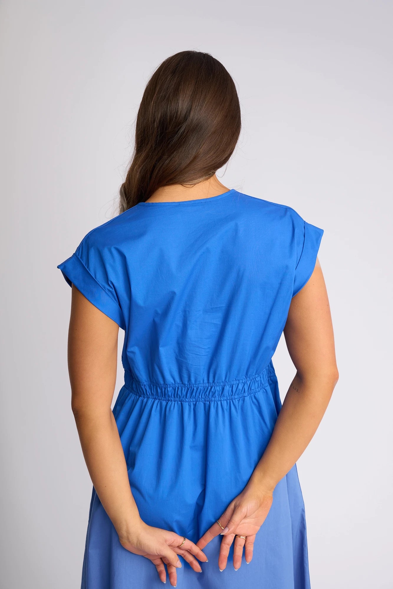A woman with long brown hair stands facing away from the camera, hands clasped behind her back, wearing the Vassalli Emmie Dress in Cobalt. The midi dress features short sleeves and a relaxed silhouette crafted from breathable cotton, lending effortless grace to her pose.