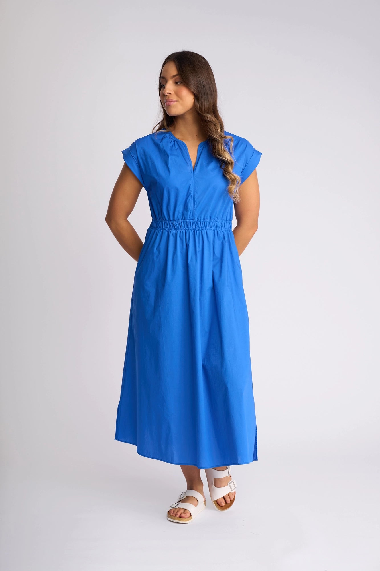 A woman stands against a plain background, wearing the Vassalli Emmie Dress in Cobalt—a bright blue, breathable cotton midi dress featuring short sleeves and an elastic waist. The relaxed silhouette complements her long hair, and she finishes the look with white sandals.