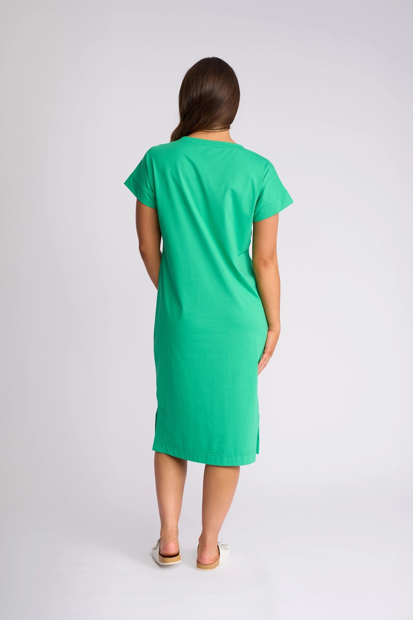 A woman with long brown hair stands facing away in a plain studio setting, wearing the Vassalli Verona Dress in Summer Green—a short-sleeve, knee-length stretch cotton dress—paired with white shoes, perfect for everyday wear.