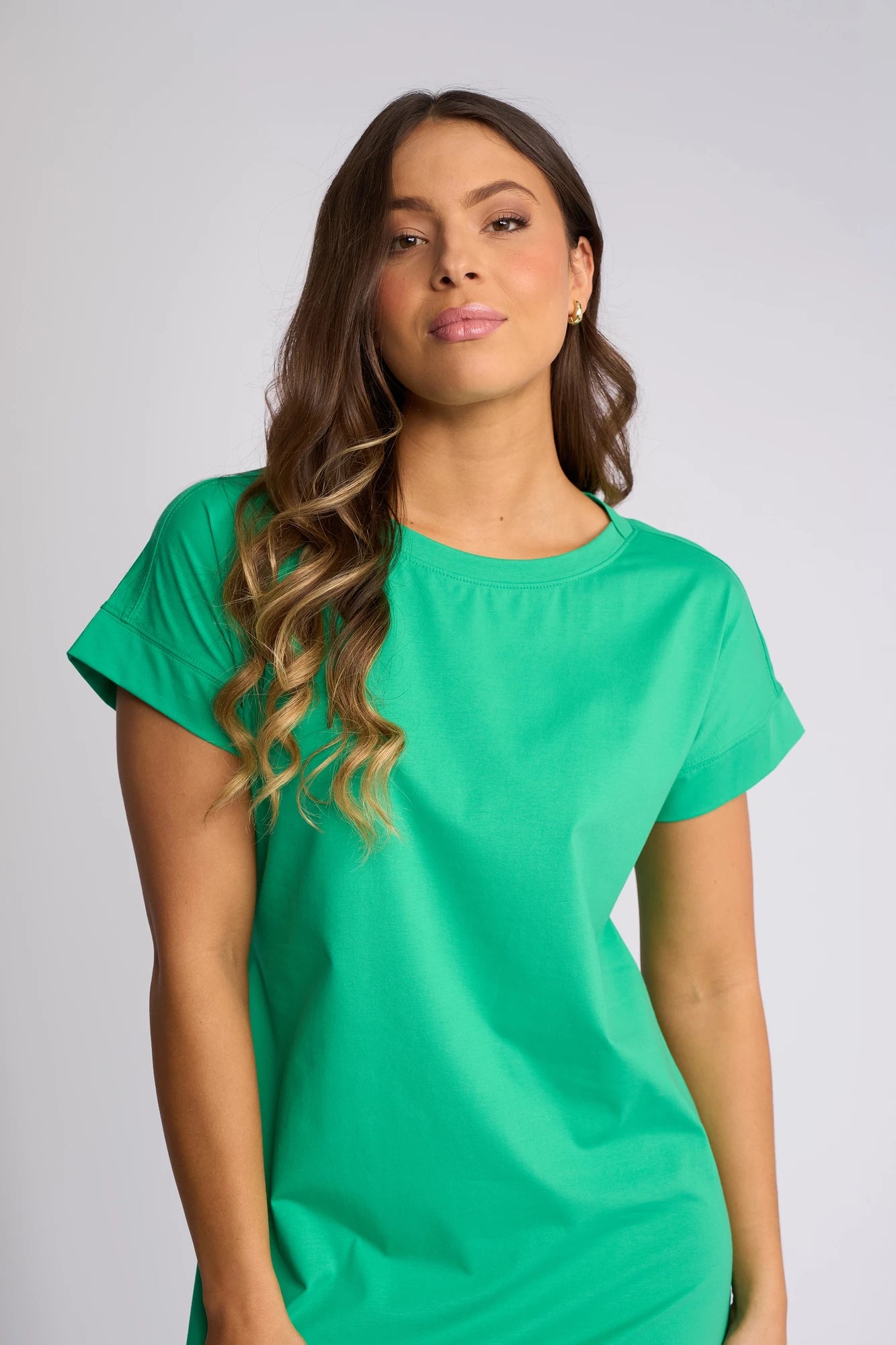 A woman with long, wavy brown hair stands confidently against a plain light gray background, wearing the Vassalli Verona Dress in Summer Green—an ideal choice for everyday wear.
