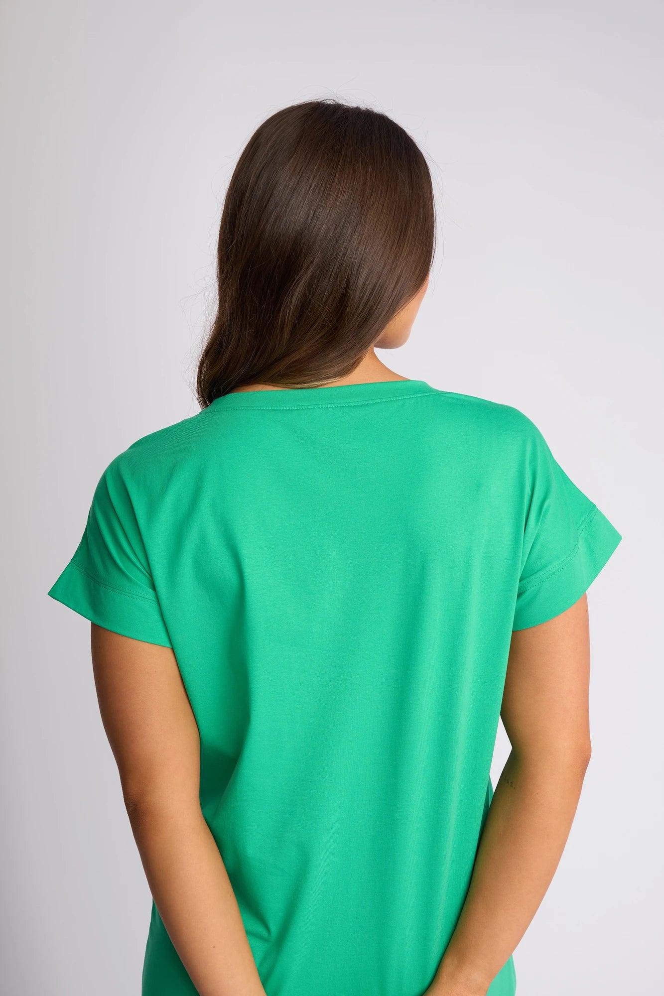 A person with long brown hair stands facing away, wearing the Vassalli Verona Dress in Summer Green—a short-sleeved stretch cotton dress—against a plain light background. Their arms rest at their sides, and their head is slightly turned to the left, showcasing an effortless style ideal for everyday wear.