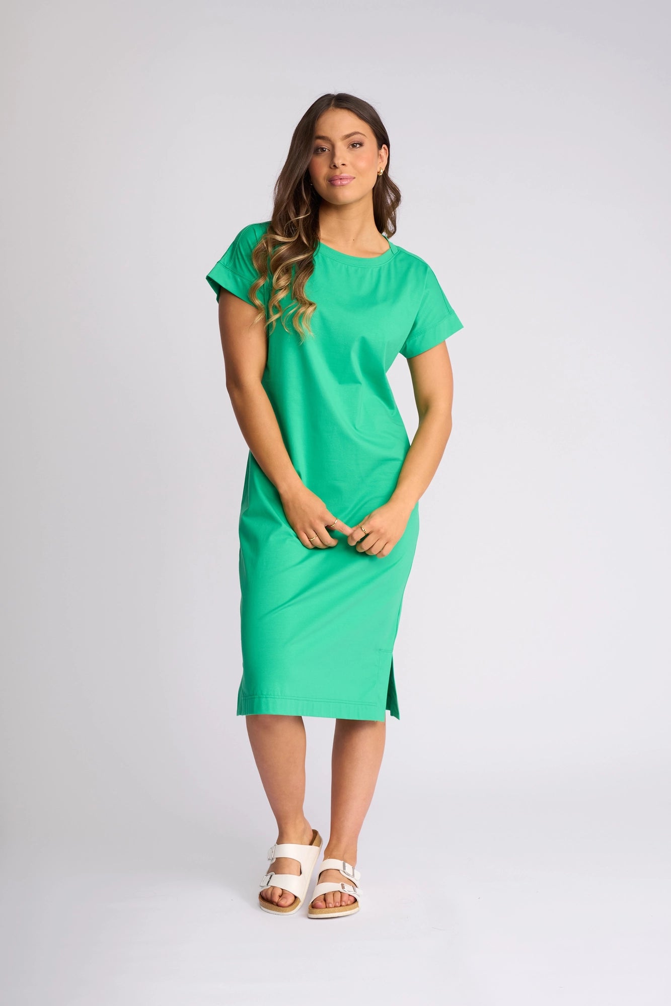 A woman with long, wavy brown hair stands against a plain light background wearing the Vassalli Verona Dress in Summer Green—a bright green, short-sleeve, stretch cotton midi dress featuring a side slit—paired with white sandals for an effortlessly stylish everyday look.