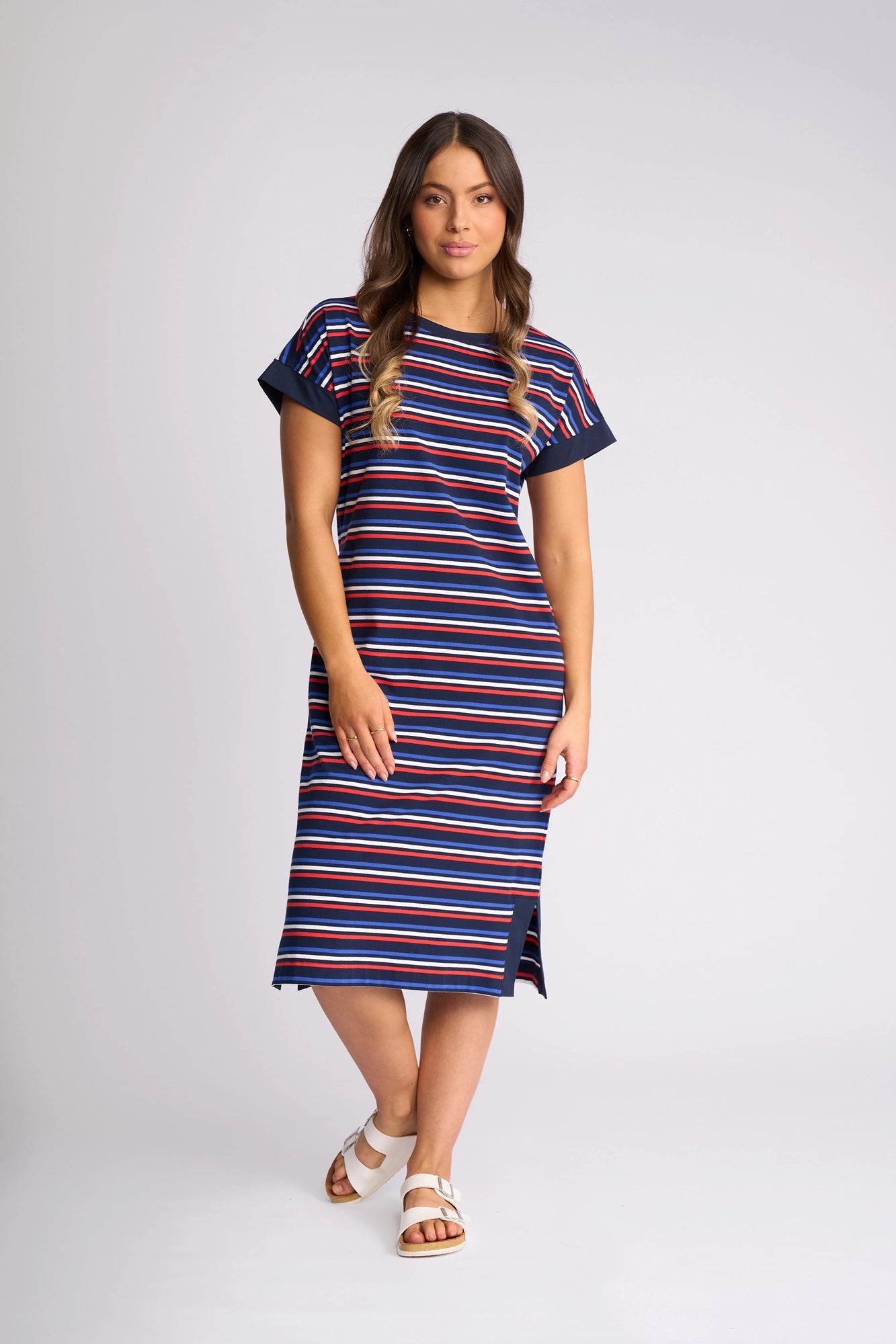 A woman stands against a plain background wearing the Vassalli Verona Dress – Mixed Stripe, a knee-length, short-sleeved cotton T-shirt dress featuring bold horizontal red, blue, and white stripes. With long wavy hair and white sandals, she effortlessly highlights the casual charm of this versatile style.