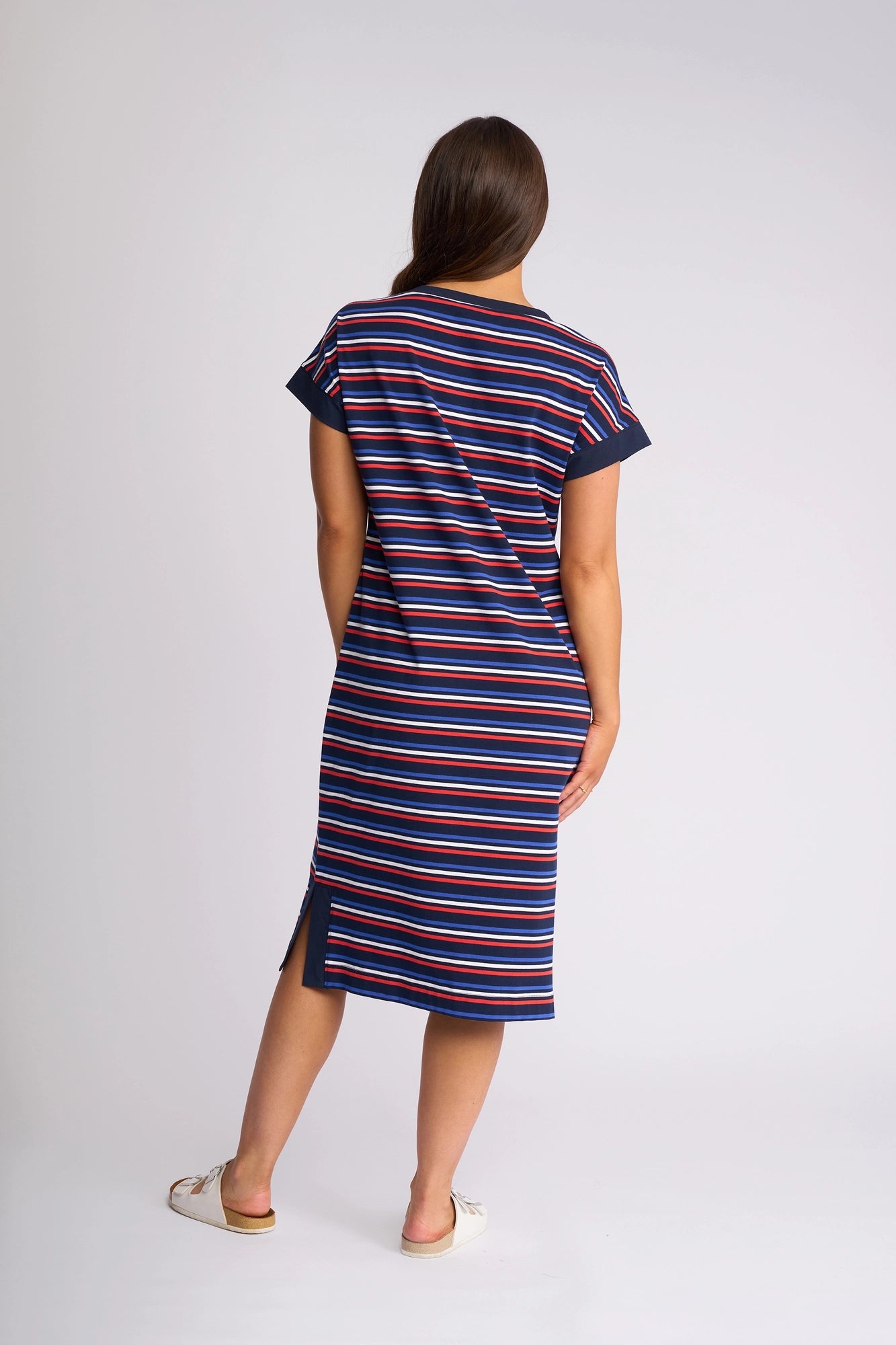 A woman stands facing away, wearing the Vassalli Verona Dress - Mixed Stripe, a knee-length, short-sleeved navy T-shirt dress featuring horizontal red, white, and blue stripes, paired with white slip-on shoes.