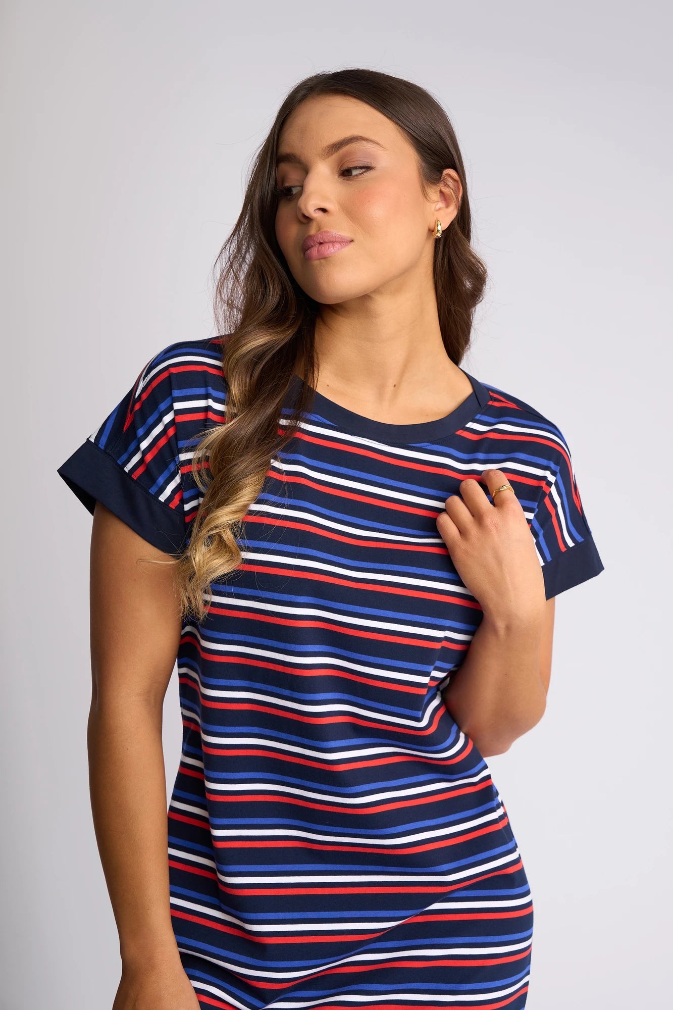 A woman stands against a light background, looking to her left with one hand touching her collar, wearing the Vassalli Verona Dress - Mixed Stripe: a short-sleeved cotton dress in navy, white, and red stripes.