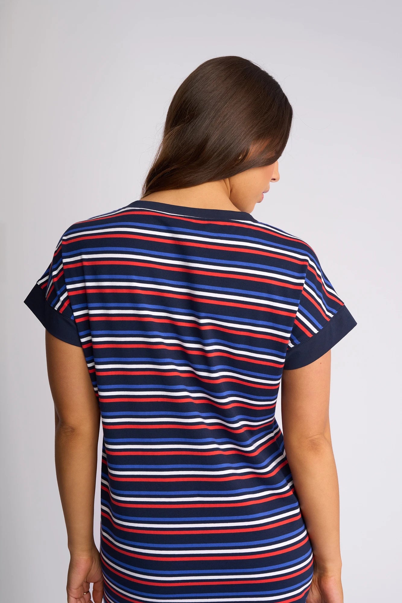 A person with long brown hair stands facing away, wearing the Vassalli Verona Dress - Mixed Stripe. The short-sleeved dress features horizontal red, white, and blue stripes. The background is plain and light-colored.