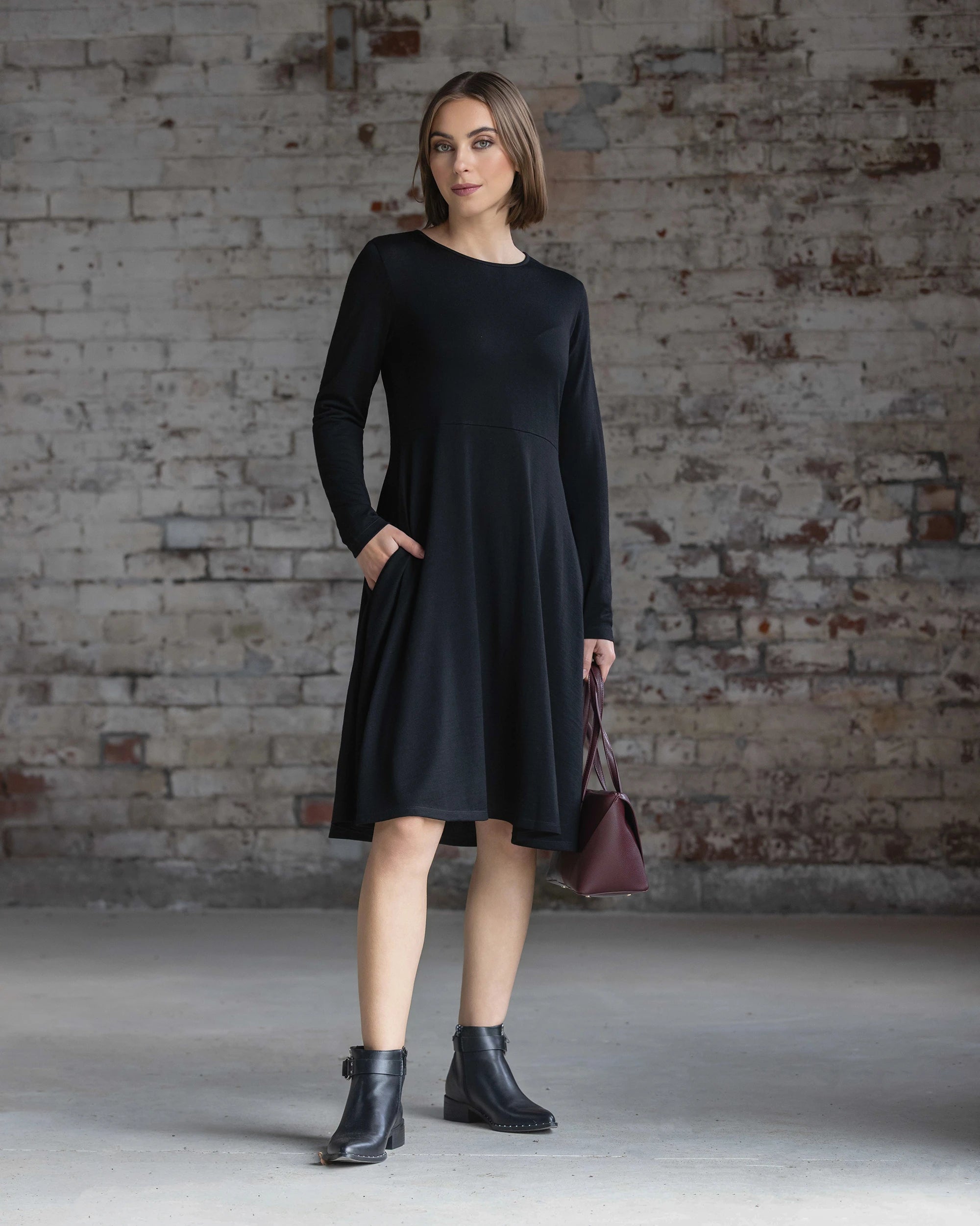 A woman with short brown hair and a neutral expression stands before a textured brick wall, wearing the Vassalli Lotus Merino Dress in black with black ankle boots and holding a burgundy handbag.
