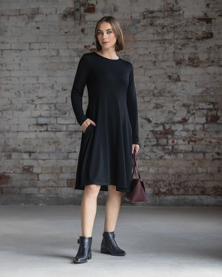 A woman with short brown hair and a neutral expression stands before a textured brick wall, wearing the Vassalli Lotus Merino Dress in black with black ankle boots and holding a burgundy handbag.