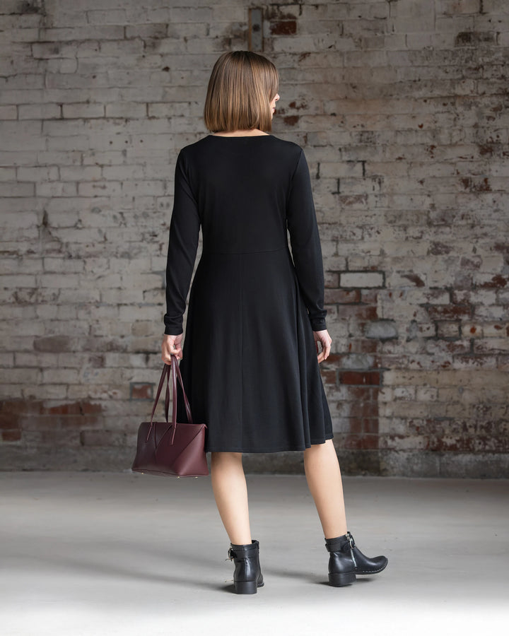 A woman wearing the Vassalli Lotus Merino Dress in black and black ankle boots faces a white brick wall, holding a maroon handbag in her left hand. Her straight hair is cut to shoulder length.