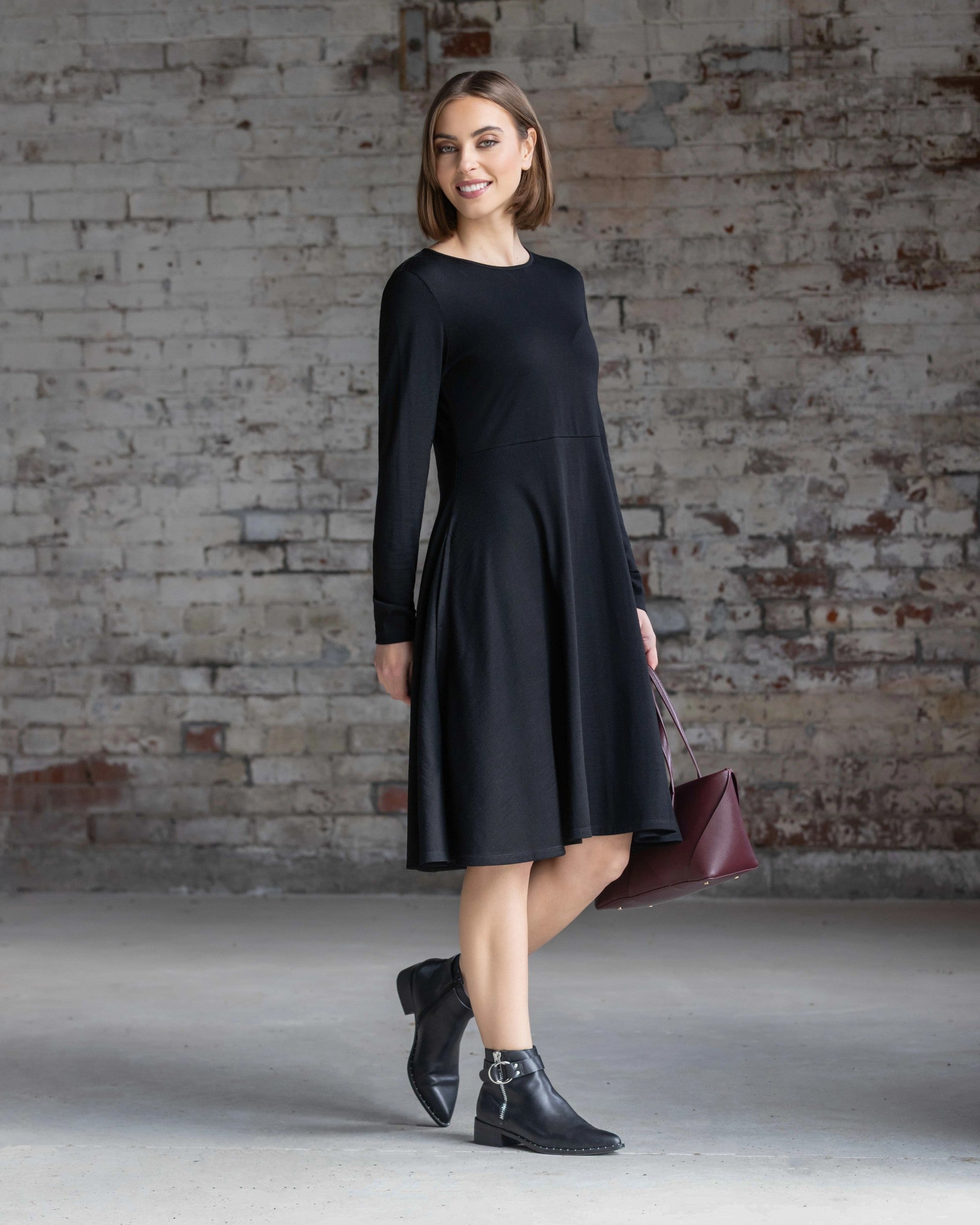 A woman wearing the Vassalli Lotus Merino Dress in black with black ankle boots and a maroon handbag stands smiling in front of a weathered white brick wall.