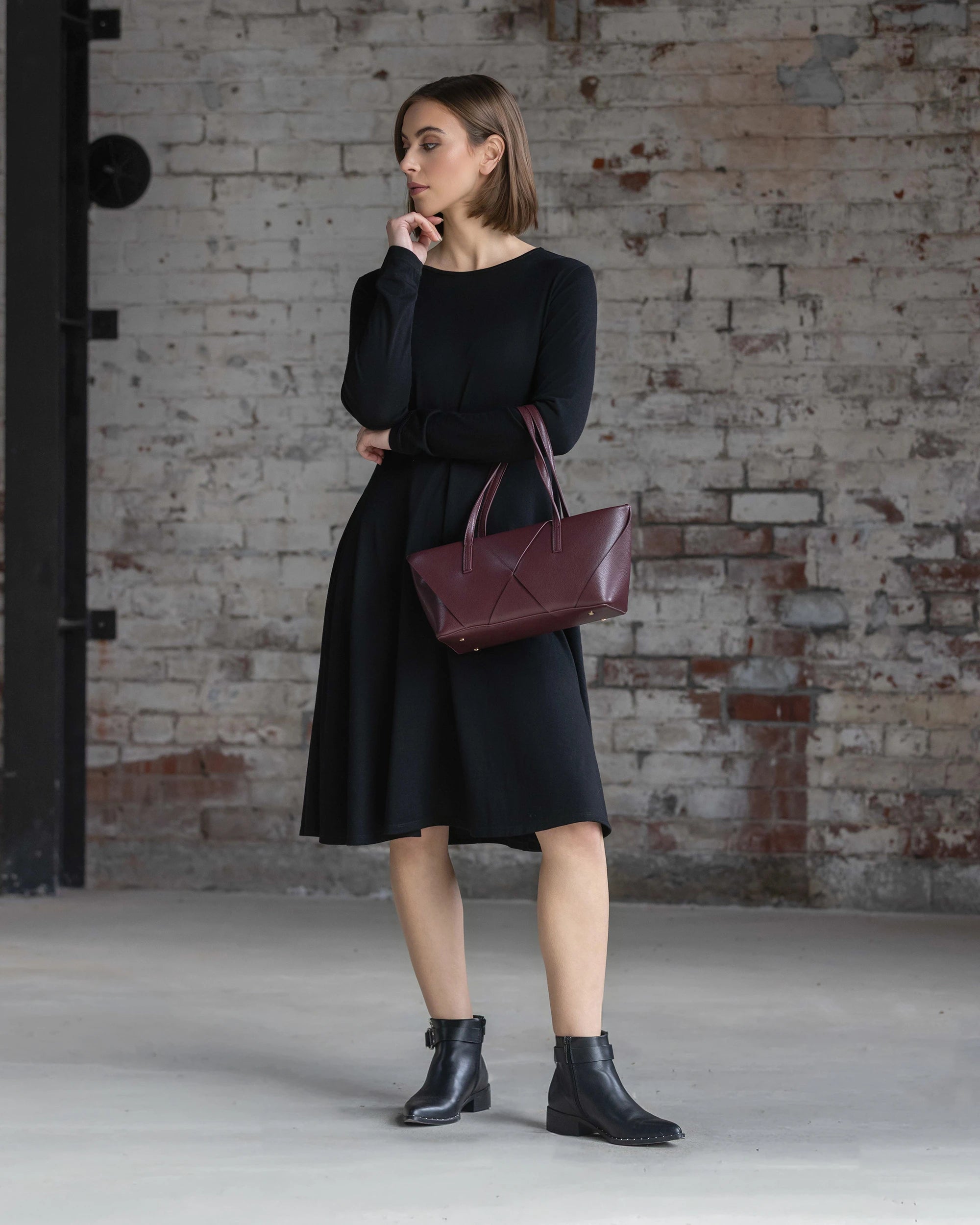 A woman wearing the Vassalli Lotus Merino Dress in black and black ankle boots stands indoors by a weathered brick wall, holding a maroon handbag and gazing thoughtfully to the side.