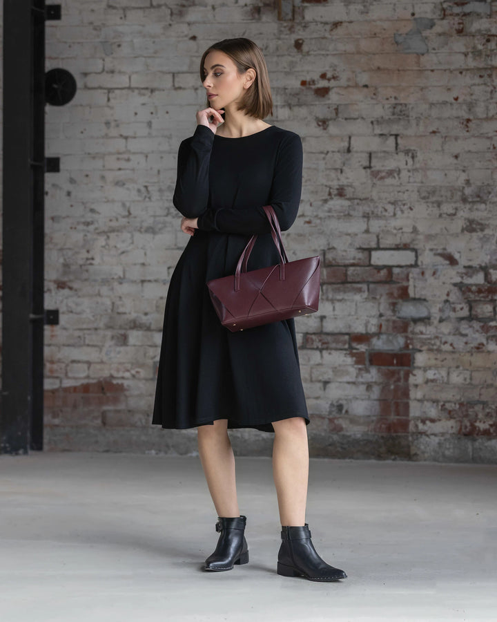 A woman wearing the Vassalli Lotus Merino Dress in black and black ankle boots stands indoors by a weathered brick wall, holding a maroon handbag and gazing thoughtfully to the side.