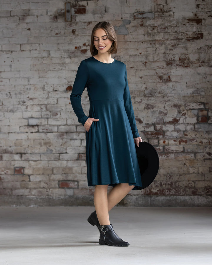 A woman with short brown hair smiles against a weathered brick wall, wearing the Vassalli Lotus Merino Dress - Dragonfly, paired with black ankle boots and a black wide-brim hat.