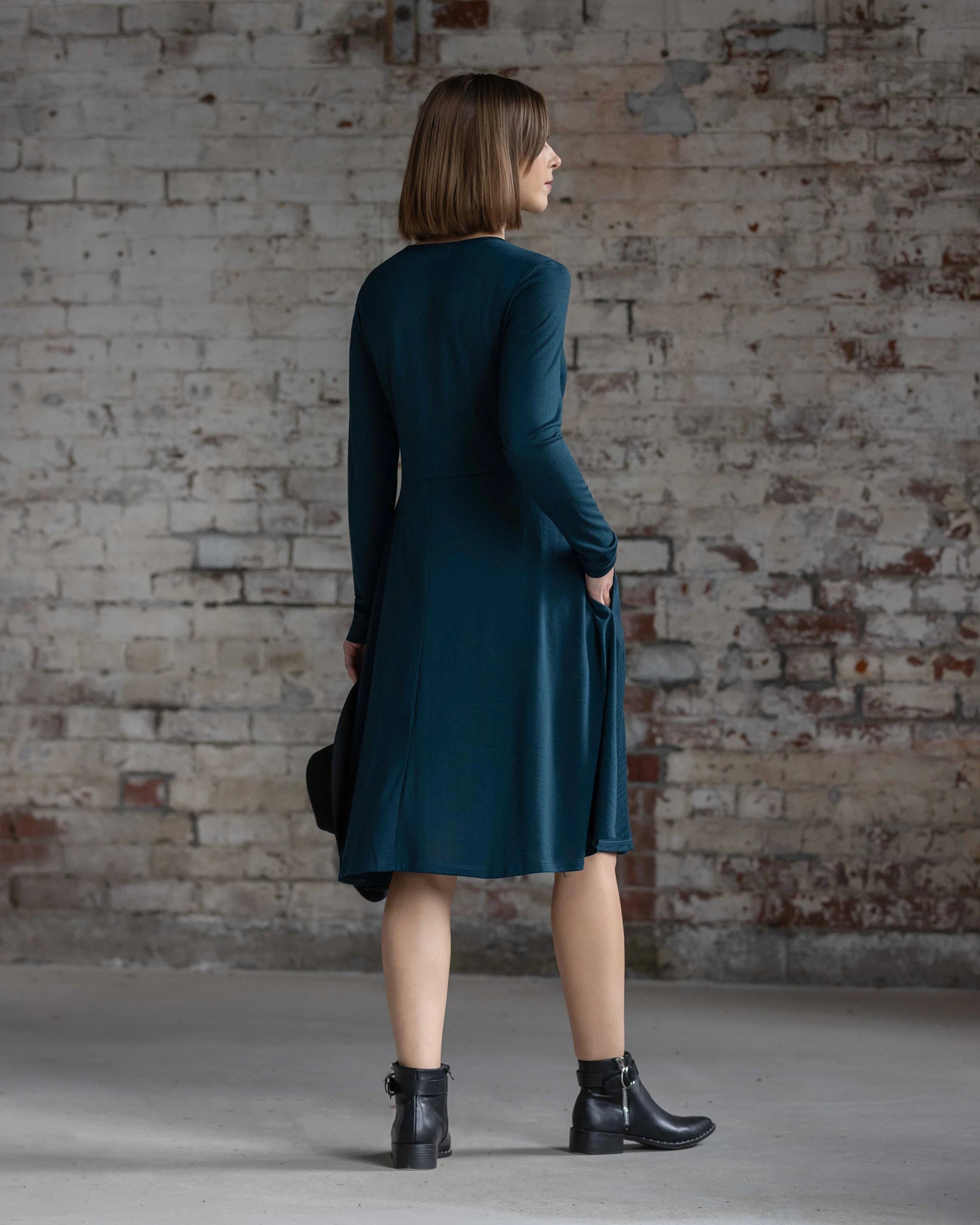 A woman with straight brown hair stands with her back to the camera, in front of a weathered brick wall, wearing the Vassalli Lotus Merino Dress - Dragonfly and black ankle boots.