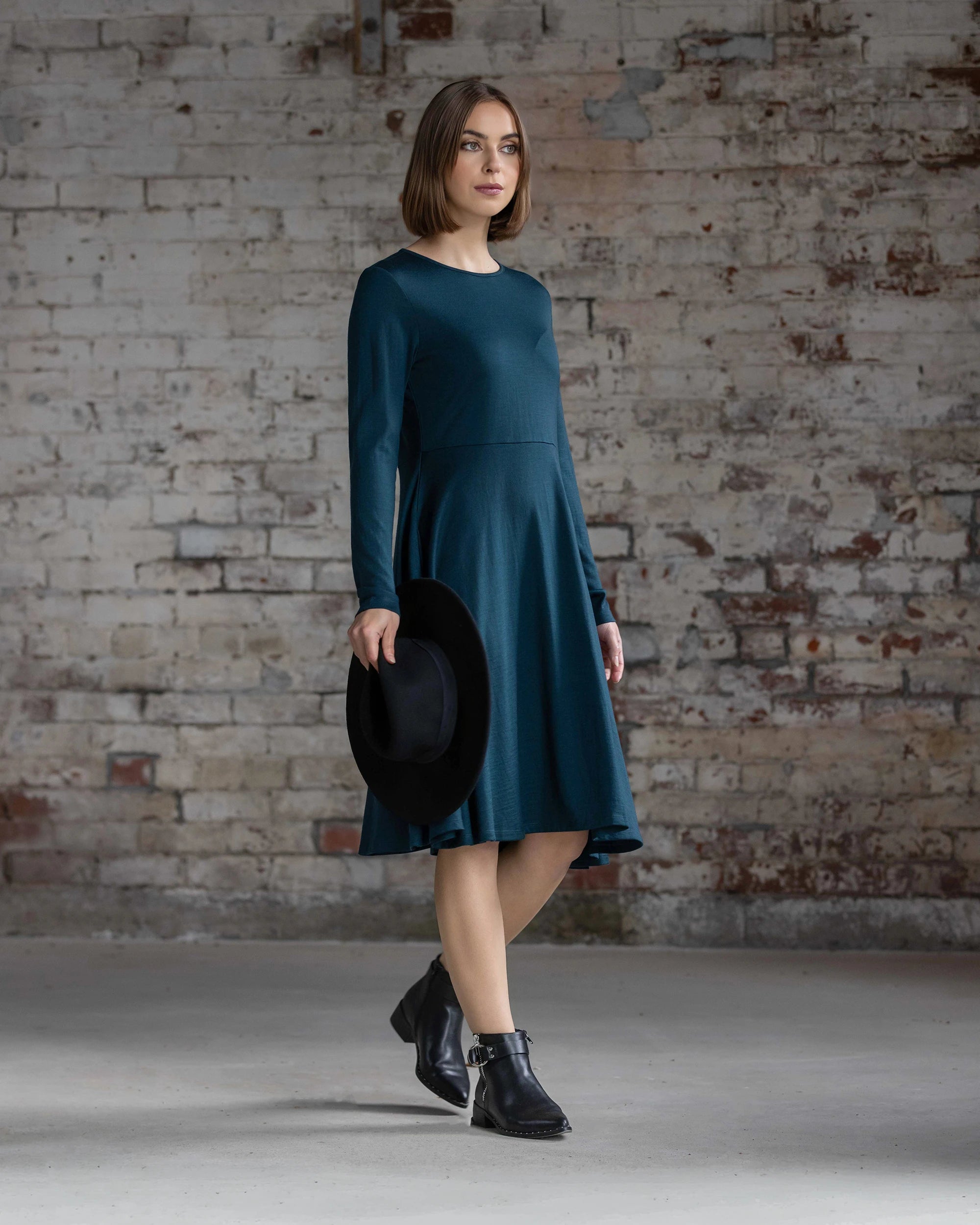 A woman in the Vassalli Lotus Merino Dress - Dragonfly, a teal knee-length piece with a slim fit, stands by a weathered brick wall wearing black ankle boots, holding a black hat and gazing to the side.