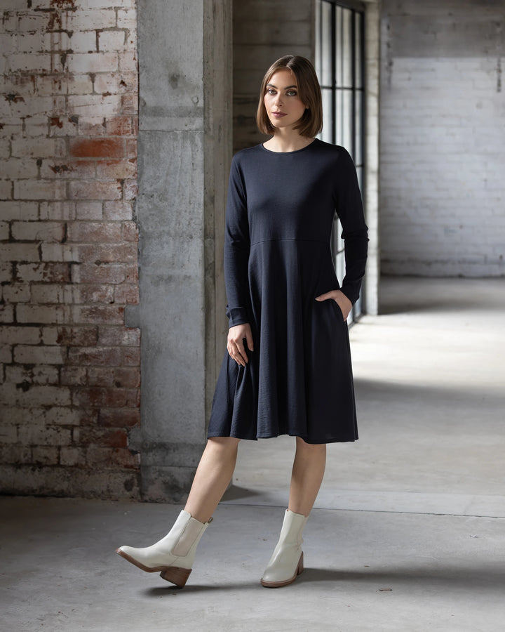 A woman wears the Vassalli Lotus Merino Dress in Midnight with white ankle boots, standing in an industrial-style room with white brick walls and large windows. She has a neutral expression and one hand in her dress pocket.
