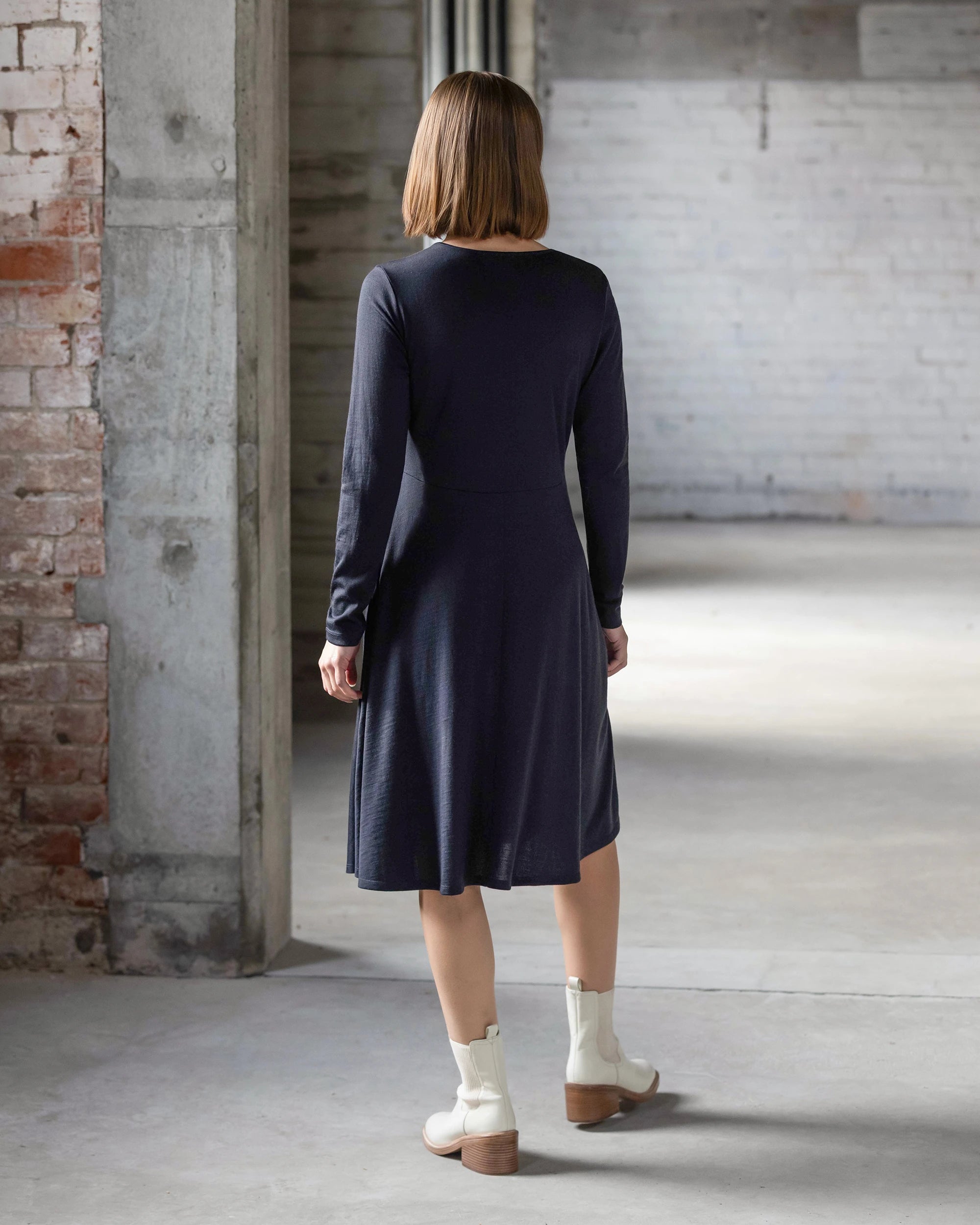 A woman with chin-length brown hair stands indoors, facing away, wearing the Vassalli Lotus Merino Dress in Midnight and off-white ankle boots in a spacious room with exposed brick and concrete walls.