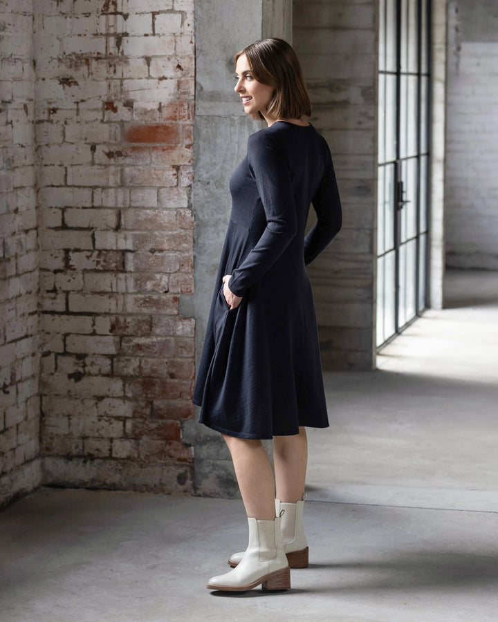 A woman wears the Vassalli Lotus Merino Dress in Midnight, a long-sleeve, knee-length swing dress, paired with white ankle boots. She stands indoors by a brick wall, smiling amid large windows and an industrial minimalist setting.