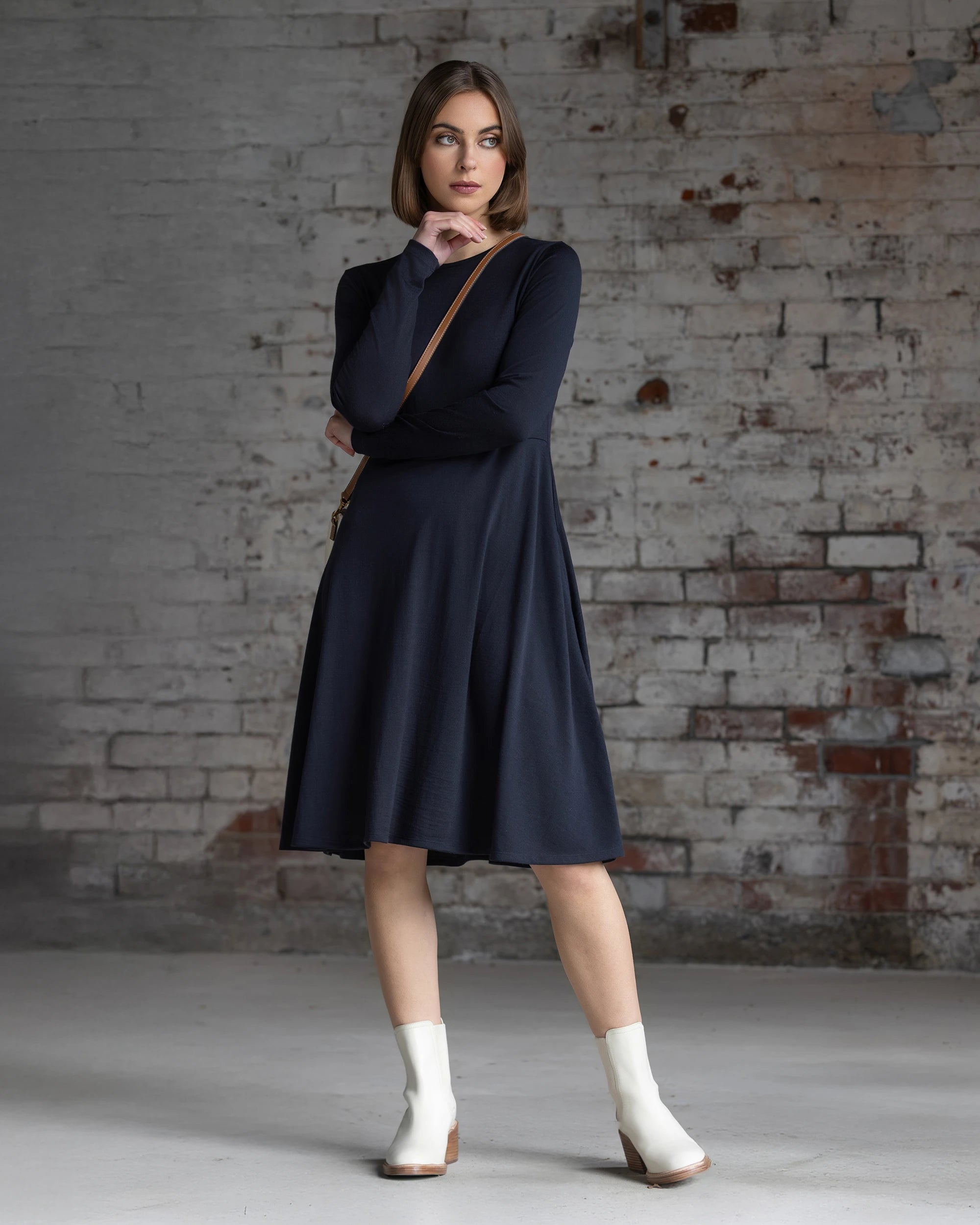 A woman in the Vassalli Lotus Merino Dress - Midnight, with long sleeves and a swing-style knee-length skirt, pairs it with white ankle boots as she poses by a worn brick wall, one hand on her chin and the other on her arm.