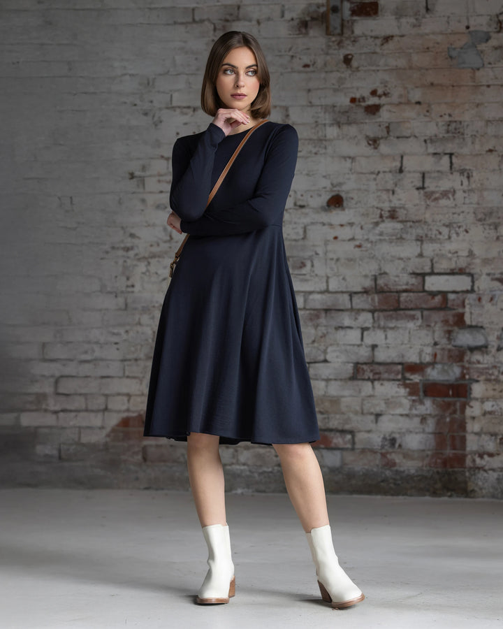 A woman in the Vassalli Lotus Merino Dress - Midnight, with long sleeves and a swing-style knee-length skirt, pairs it with white ankle boots as she poses by a worn brick wall, one hand on her chin and the other on her arm.