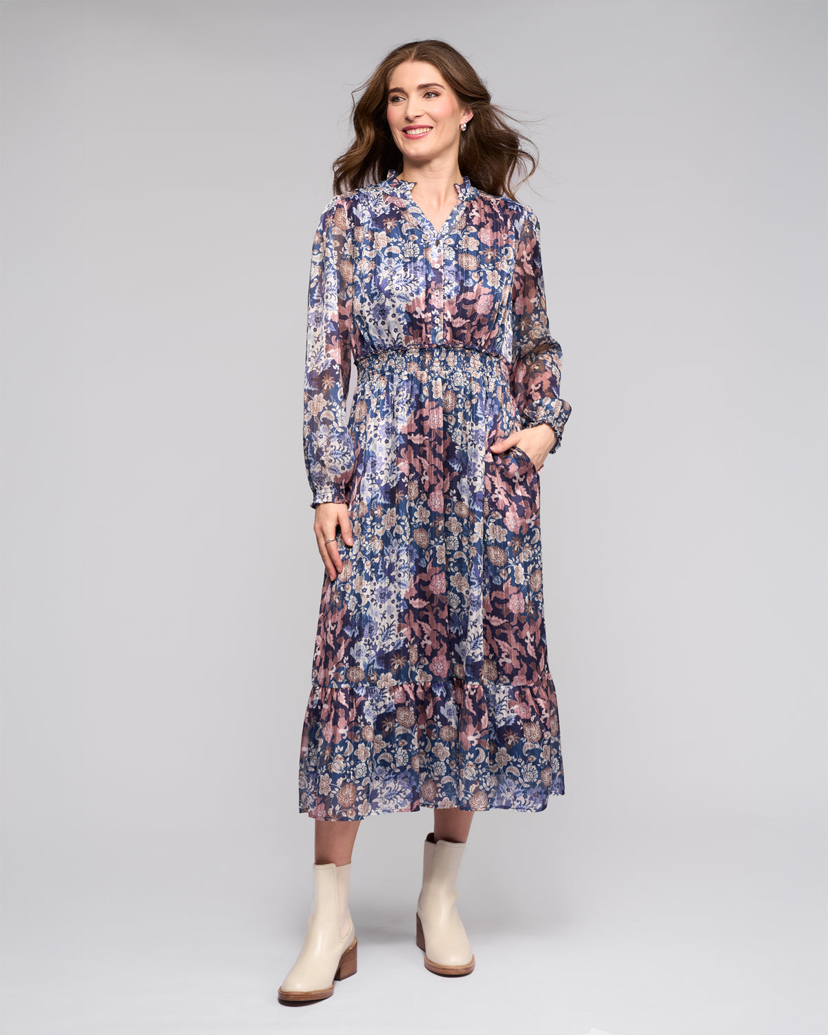 A woman smiles in the Jane Dress - Rhea by Vassalli, a floral long-sleeve dress with pockets, blue and pink hues, and a tiered skirt. She pairs it with light ankle boots against a plain light gray background.