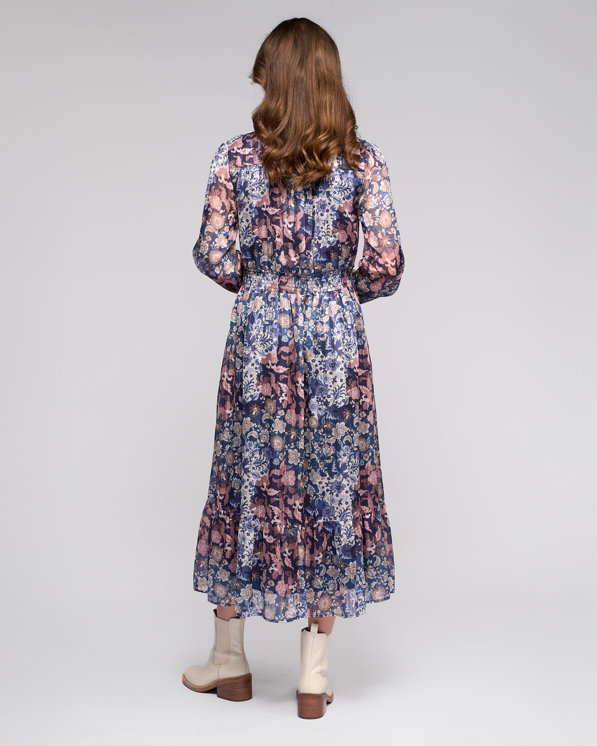 A woman with long brown hair stands facing away, wearing the Vassalli Jane Dress - Rhea—a floral long-sleeve, tiered blue and purple dress—paired with light beige ankle boots, against a plain light background.
