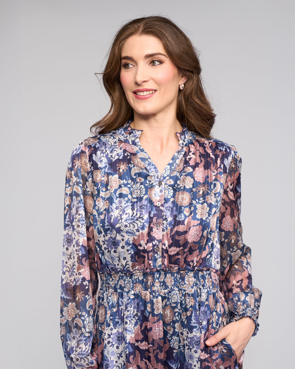 A woman with wavy brown hair wears the Vassalli Jane Dress - Rhea, a floral long-sleeve tiered dress in blue and pink, standing against a plain light gray background and smiling while looking to the side.