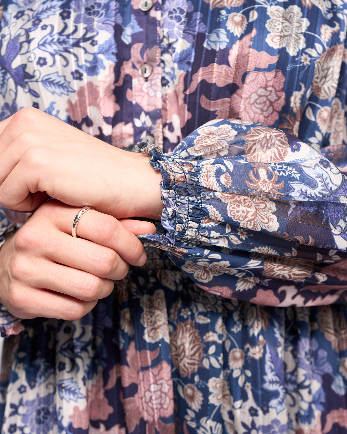A close-up of a person adjusting the sleeve of the Vassalli Jane Dress - Rhea, which features a floral print in purple, pink, white, and blue. They are also wearing a simple silver ring on their left hand.