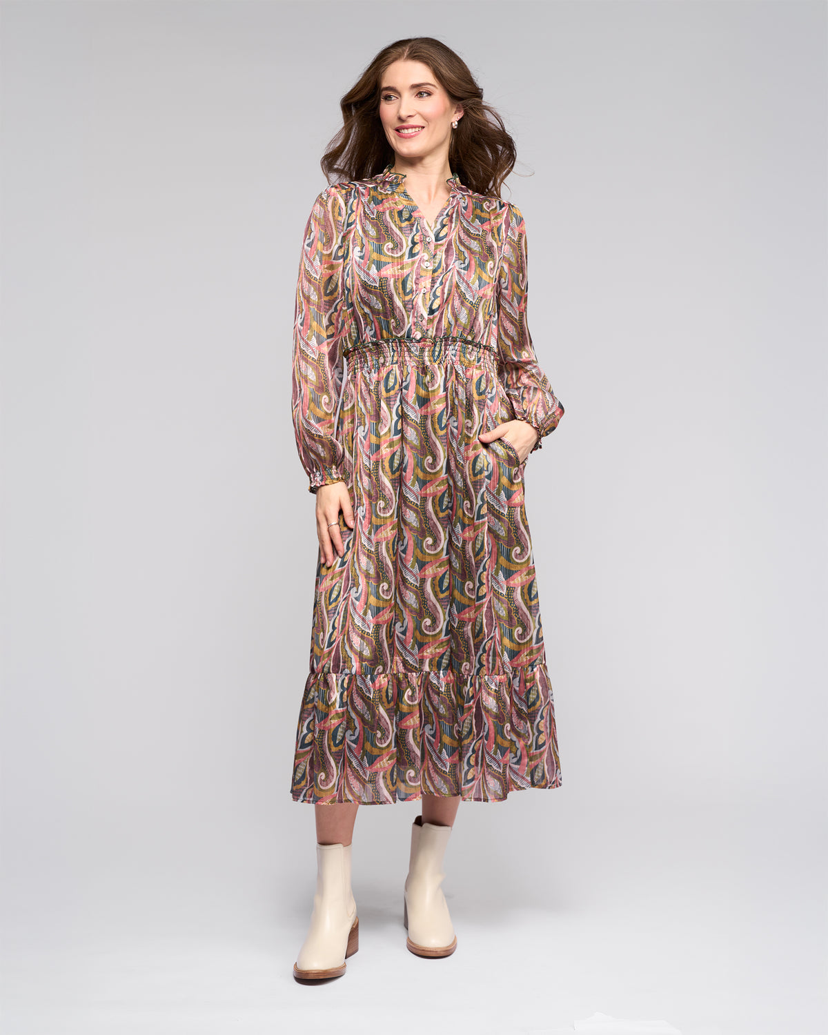A woman smiles against a plain background wearing the Vassalli Jane Dress - Sable, an ankle-length, long-sleeve dress with a colorful paisley pattern, paired with light-colored ankle boots and one hand in her pocket.