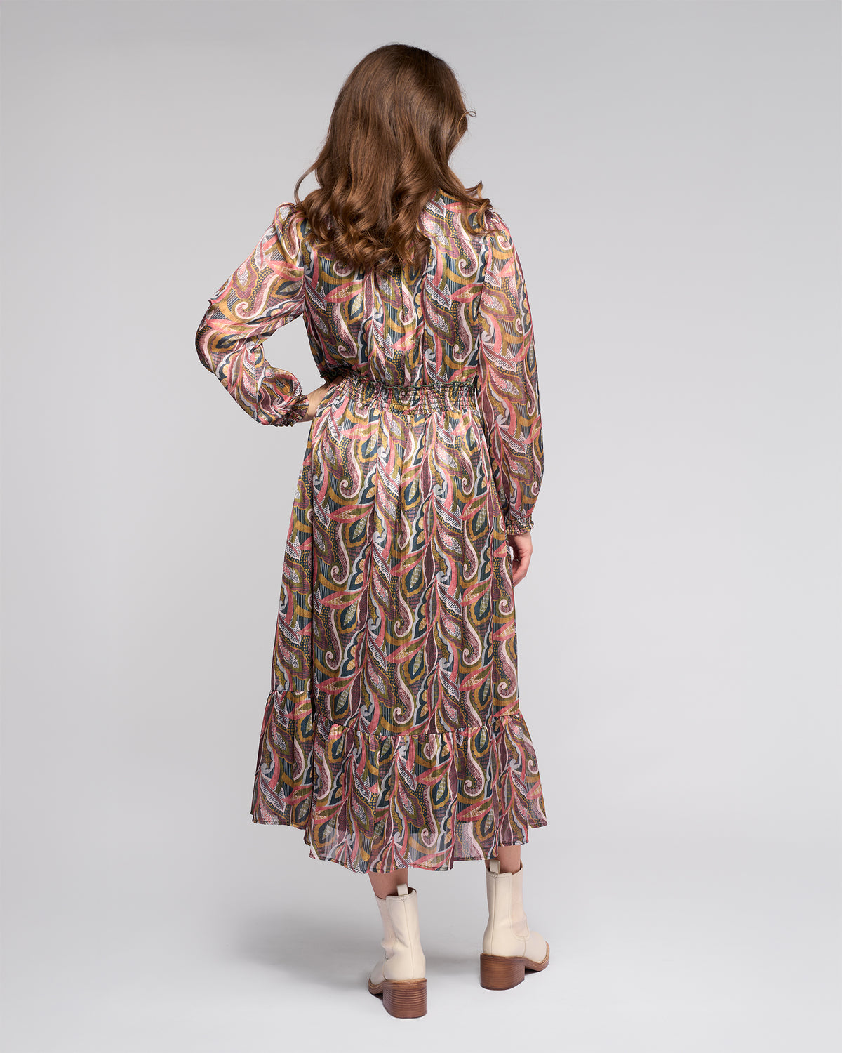 A woman with wavy brown hair, seen from behind, wears the Vassalli Jane Dress - Sable and beige ankle boots against a plain light gray background.
