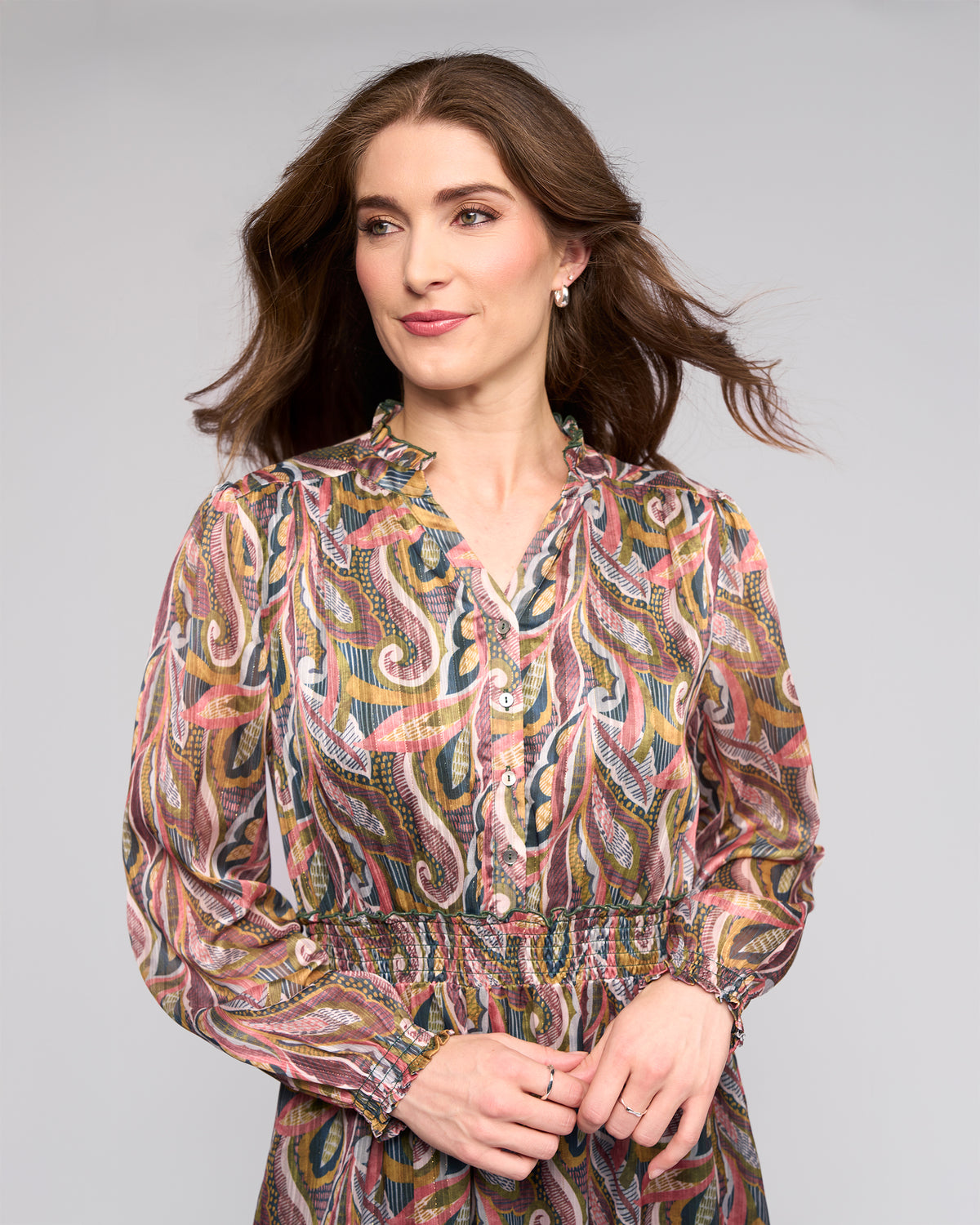 A woman with wavy brown hair wears the Vassalli Jane Dress - Sable, featuring a patterned long-sleeve design and smocked waist, standing against a plain light gray background, looking slightly left and smiling subtly.