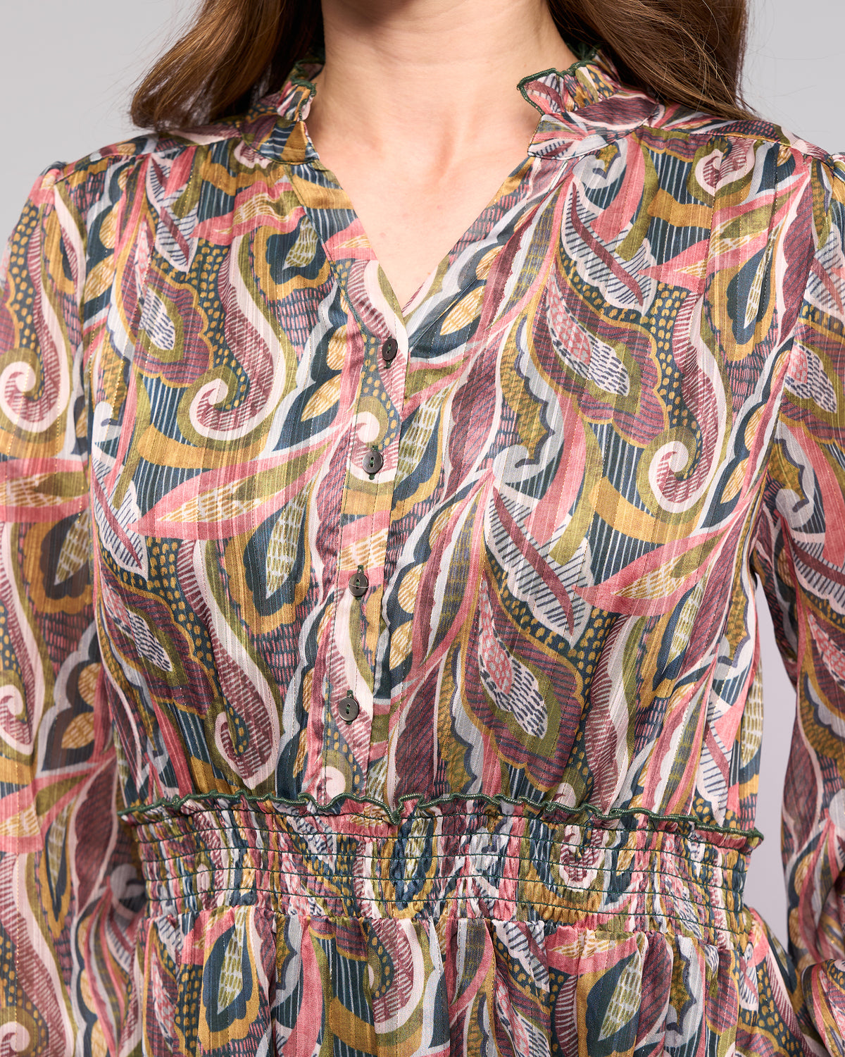 A woman wears the Vassalli Jane Dress - Sable, a colorful paisley dress with a high waist, buttoned front, long sleeves, and swirling pink, green, yellow, and white patterns.