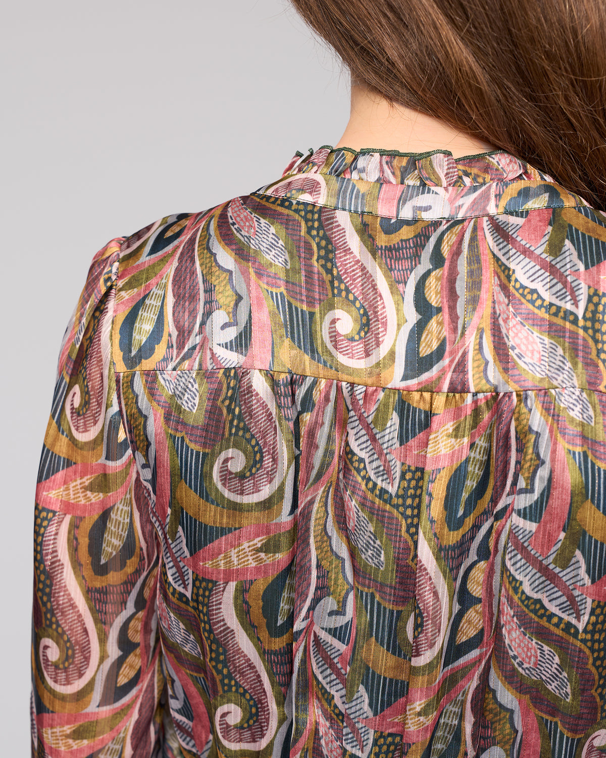 A close-up of a woman's back wearing the Vassalli Jane Dress - Sable, a sheer design with colorful swirling paisley patterns in pink, green, gold, and black. Her long brown hair drapes over one shoulder.