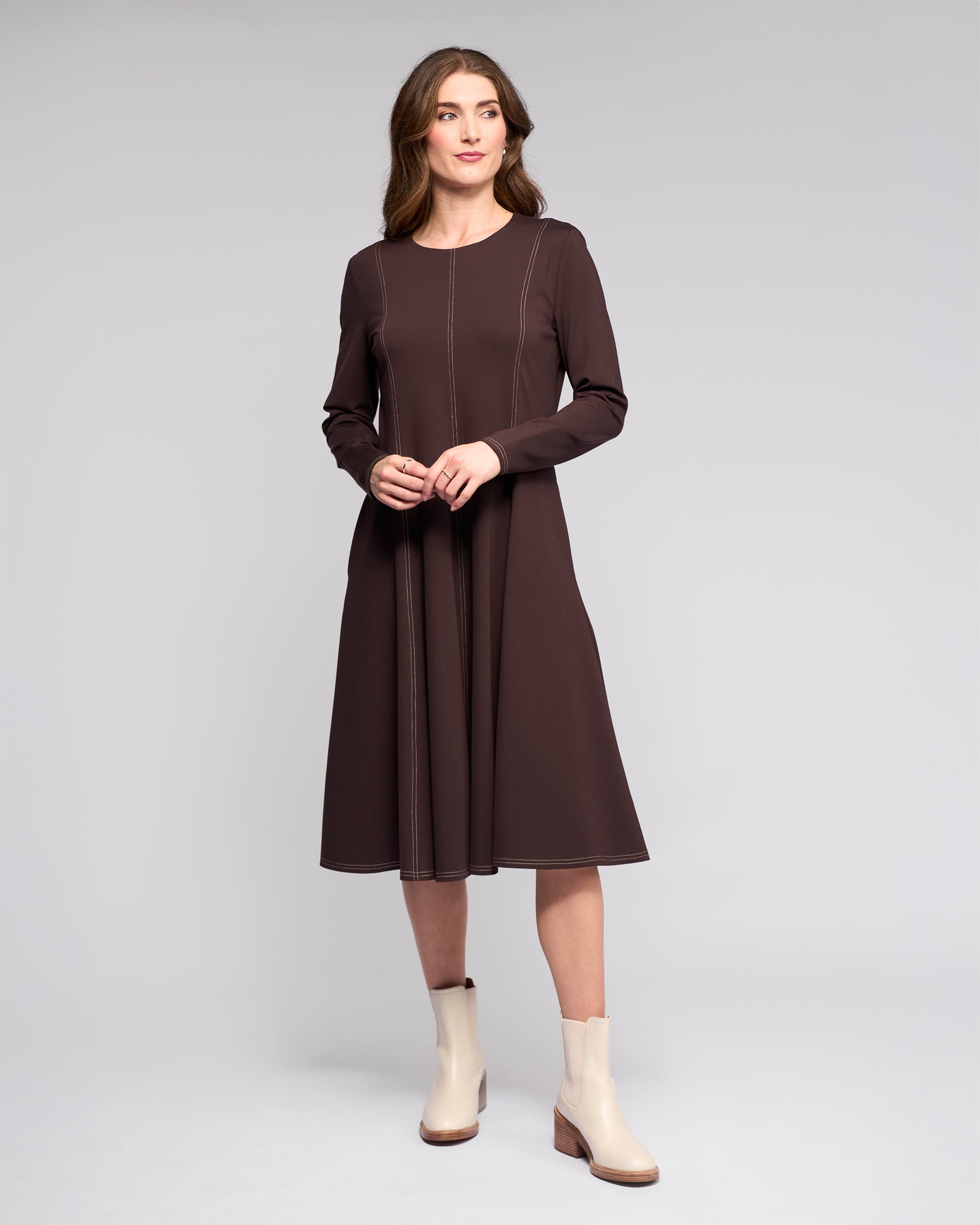 A woman stands against a light gray background wearing the Vassalli Miner Dress - Chocolate, styled with light ankle boots. She looks to the side with hands loosely clasped, highlighting the flattering silhouette of the dress.