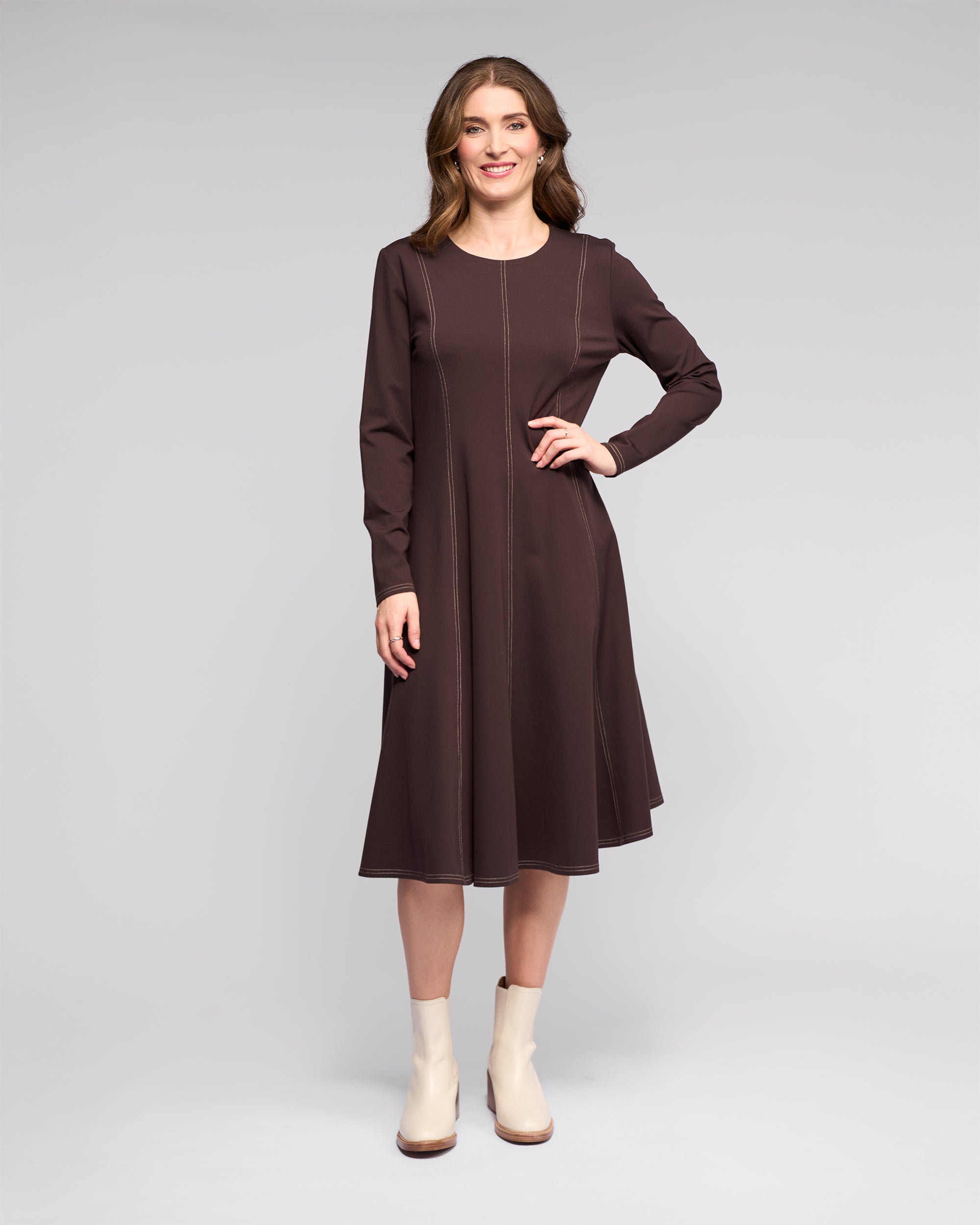 A woman faces forward, smiling in the Vassalli Miner Dress - Chocolate, featuring light stitching for a flattering silhouette, paired with cream ankle boots against a plain light gray background.