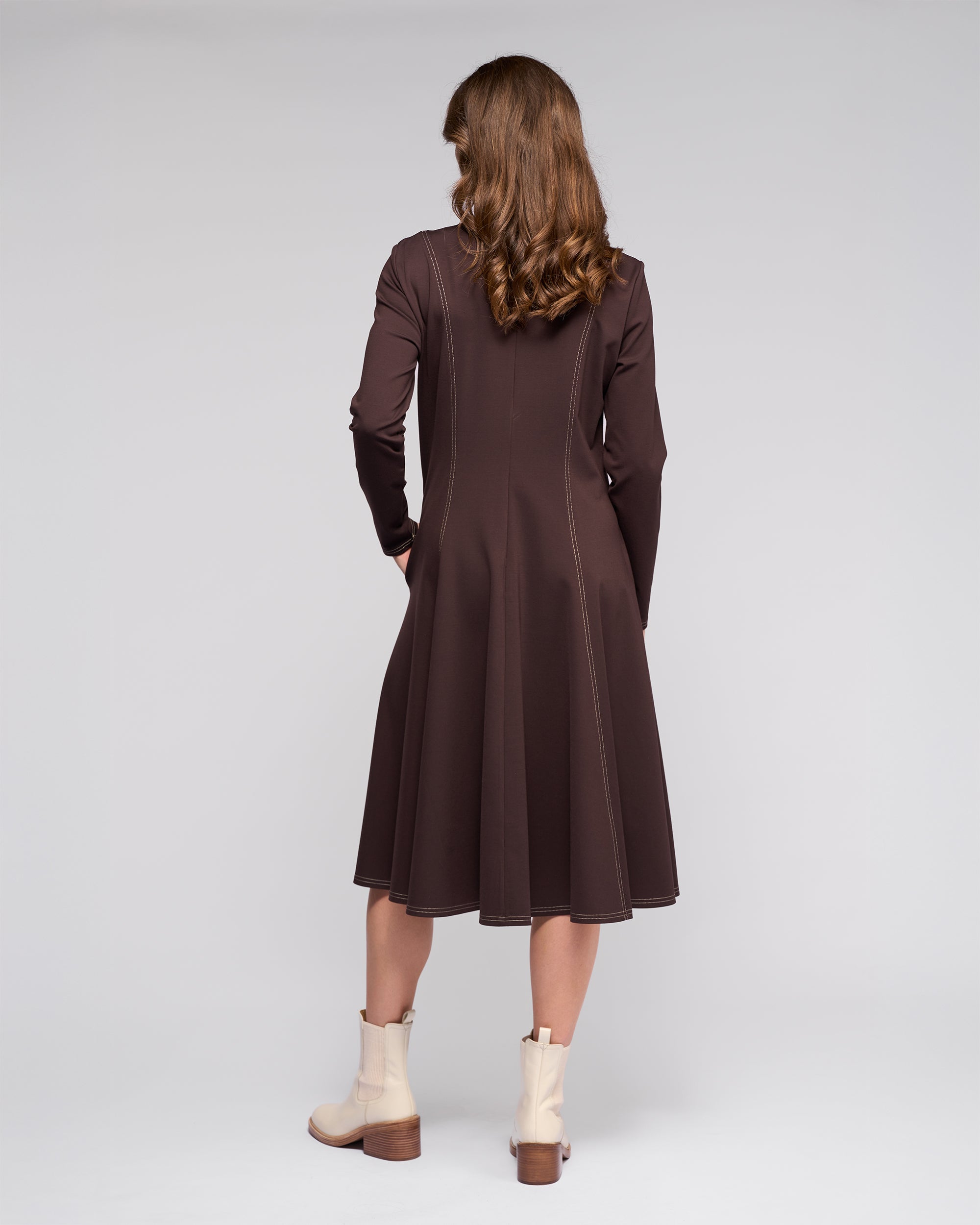 A woman with wavy brown hair stands facing away in the Vassalli Miner Dress - Chocolate, styled with light beige ankle boots against a plain light gray background.