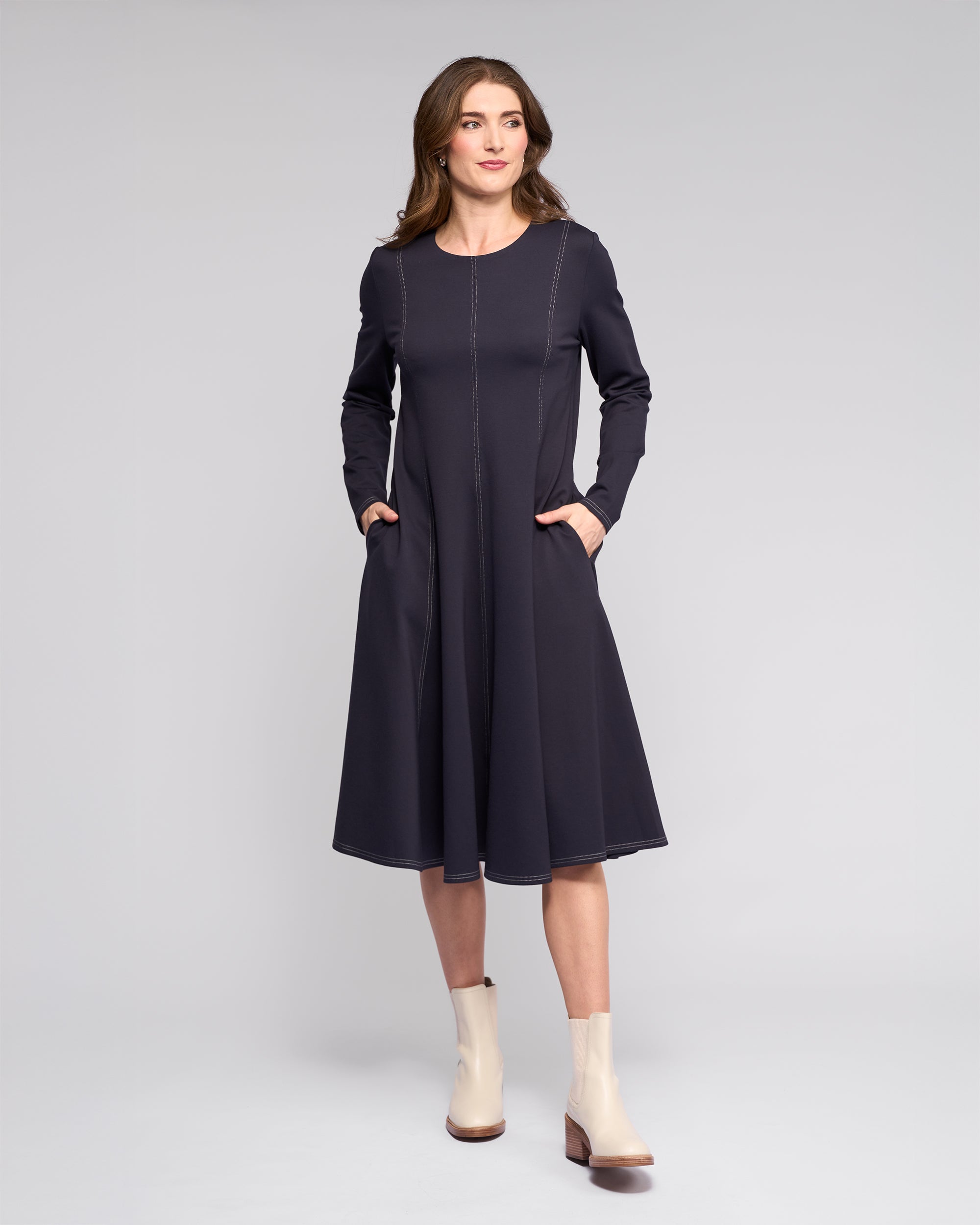 A woman stands against a light gray background wearing the Vassalli Miner Dress in Ink, a winter-weight ponti midi dress with subtle stitching, paired with cream ankle boots. She smiles softly with one hand in her pocket.