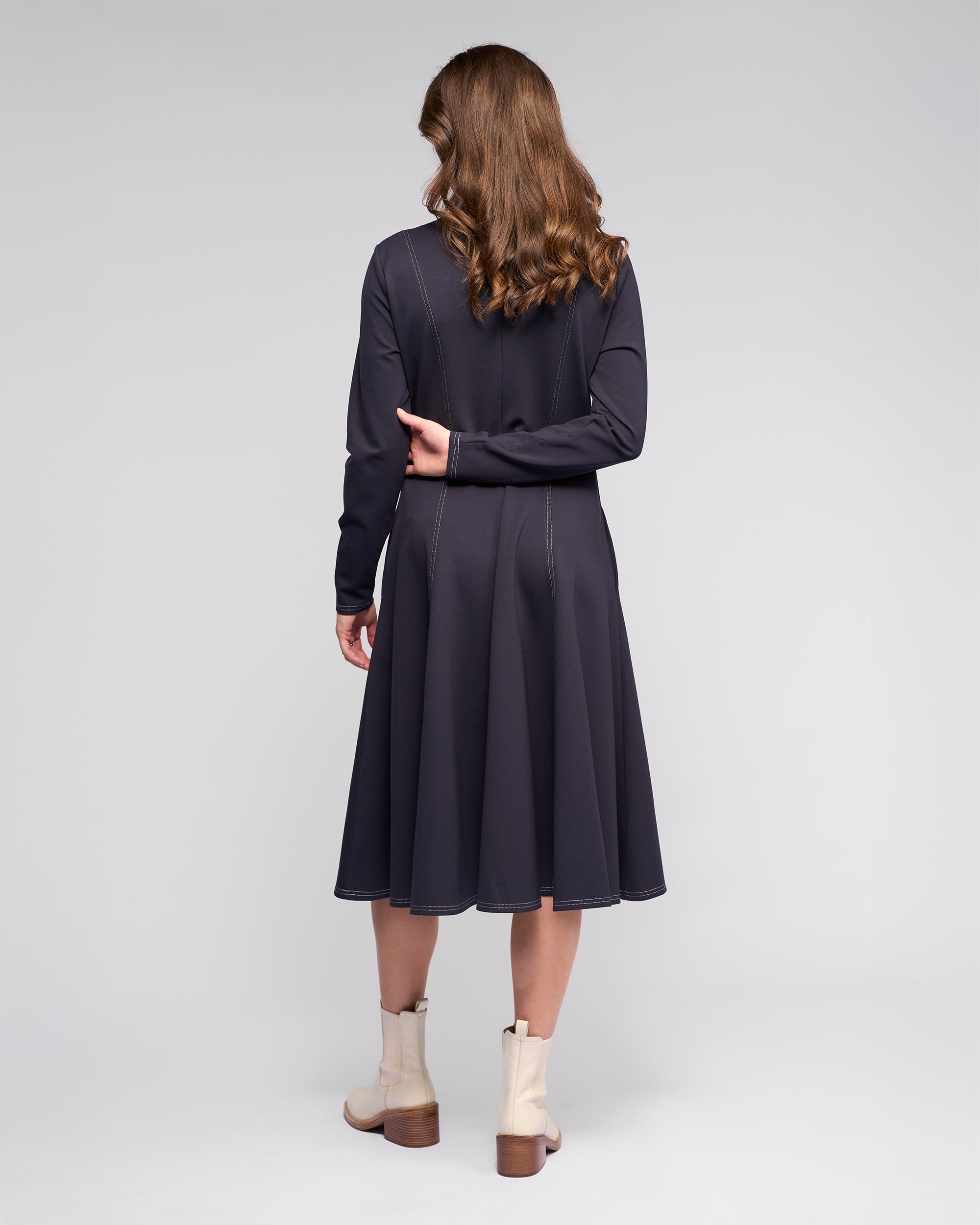A woman with wavy brown hair faces away, wearing the Vassalli Miner Dress - Ink, a deep dark navy midi in winter-weight ponti with white stitching, paired with cream ankle boots. She rests her left arm behind her back against a light gray background.