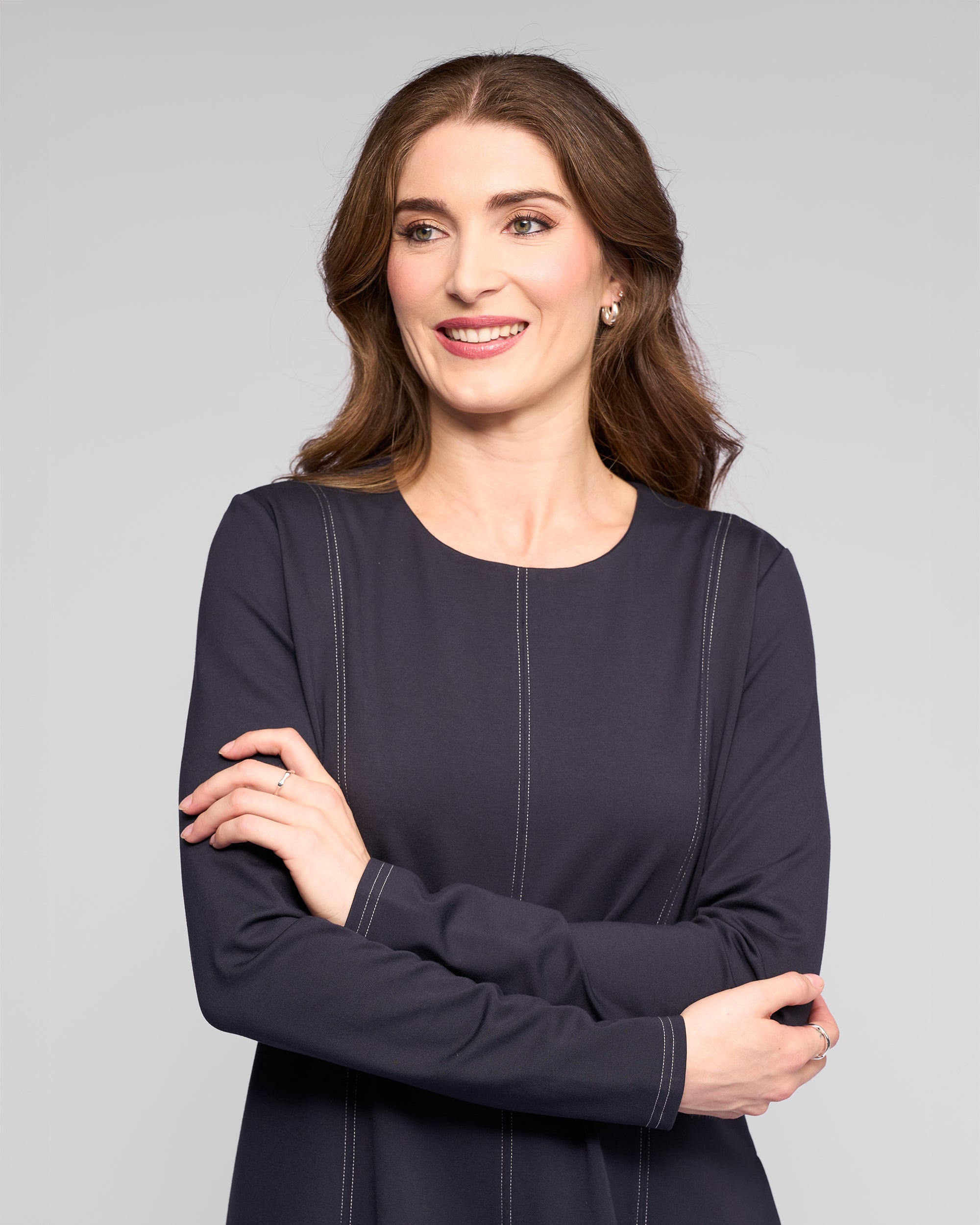 A woman with long brown hair and light skin smiles with arms crossed, wearing the Vassalli Miner Dress - Ink, a deep dark navy winter-weight ponti midi dress, against a plain light gray background.
