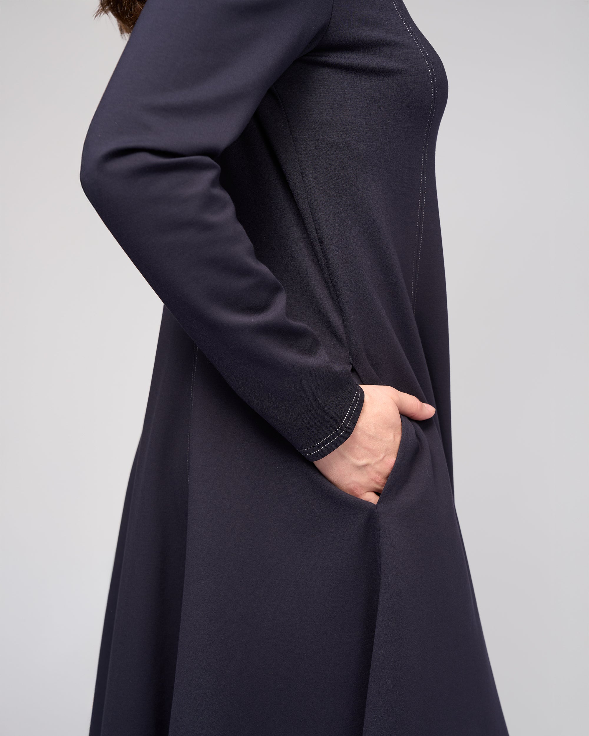 A person is shown from the side with their right hand in a side pocket, wearing the Vassalli Miner Dress - Ink, a midi-length, deep navy winter-weight ponti dress, against a plain light-colored background.