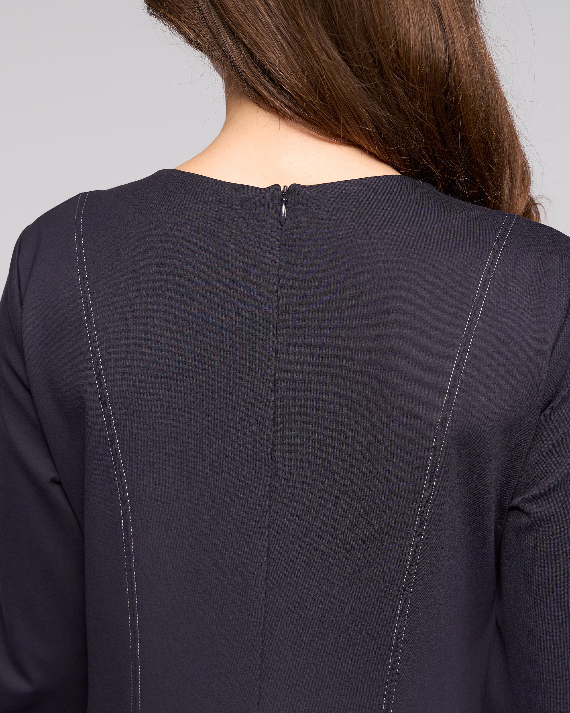 A close-up of a person with long brown hair wearing the Vassalli Miner Dress - Ink, featuring a deep navy winter-weight ponti fabric, white stitched lines, and a small button at the neckline.