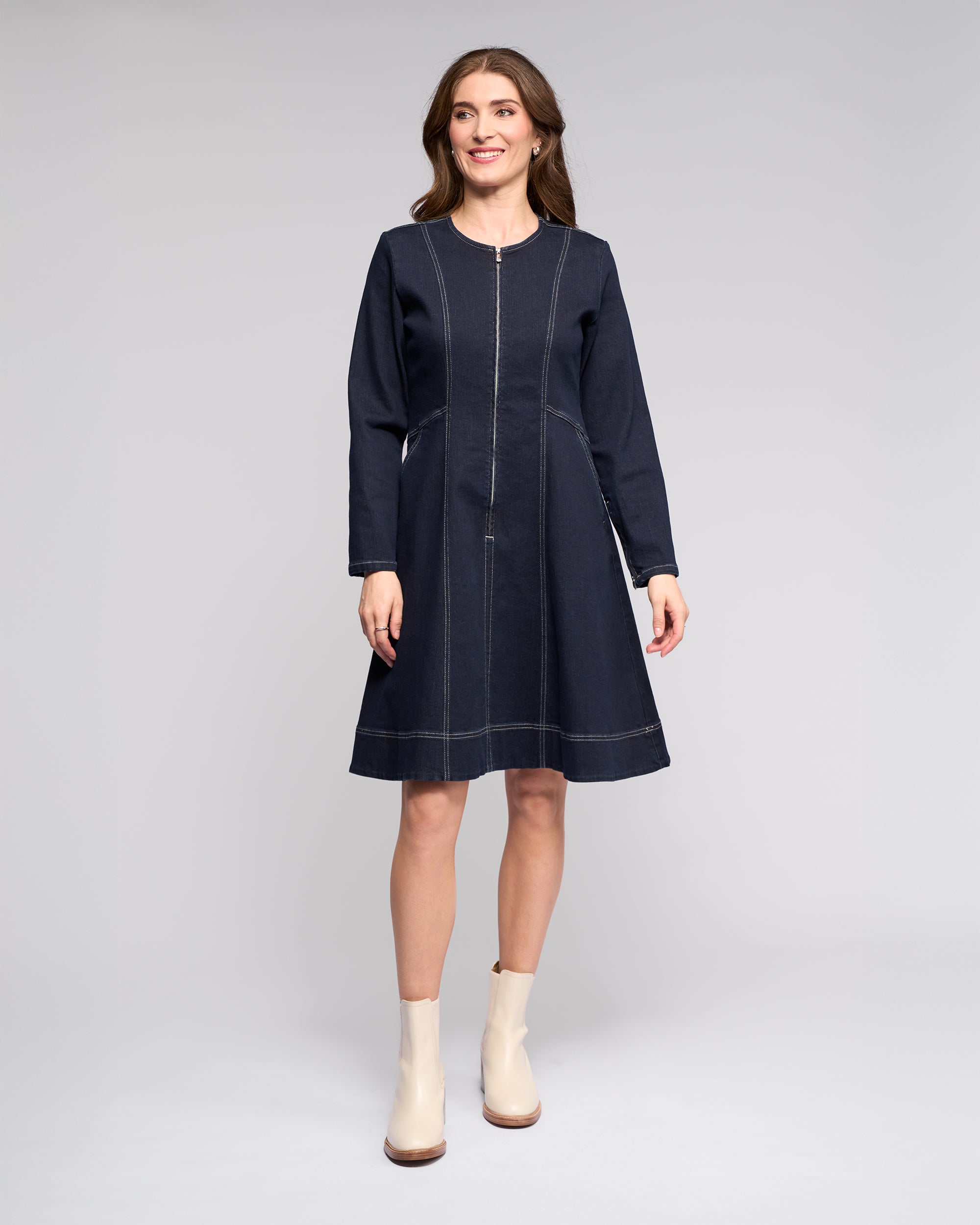 A woman smiles against a light gray background, wearing the Vassalli Sienna Dress in Ink Denim with white stitching and cream ankle boots. Her loose hair adds a relaxed touch to the chic look.