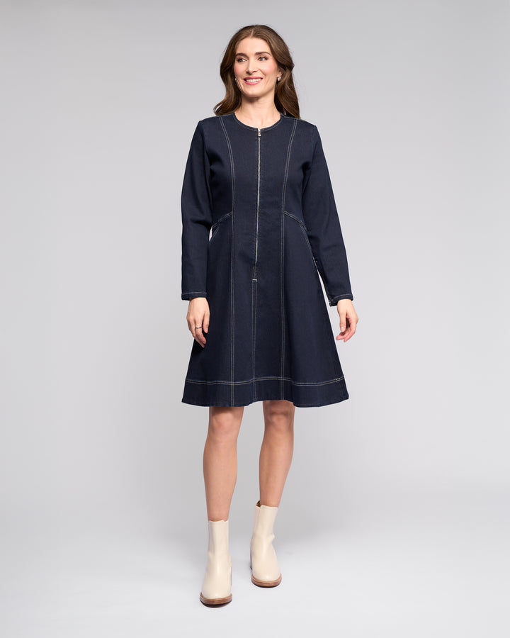 A woman smiles against a light gray background, wearing the Vassalli Sienna Dress in Ink Denim with white stitching and cream ankle boots. Her loose hair adds a relaxed touch to the chic look.