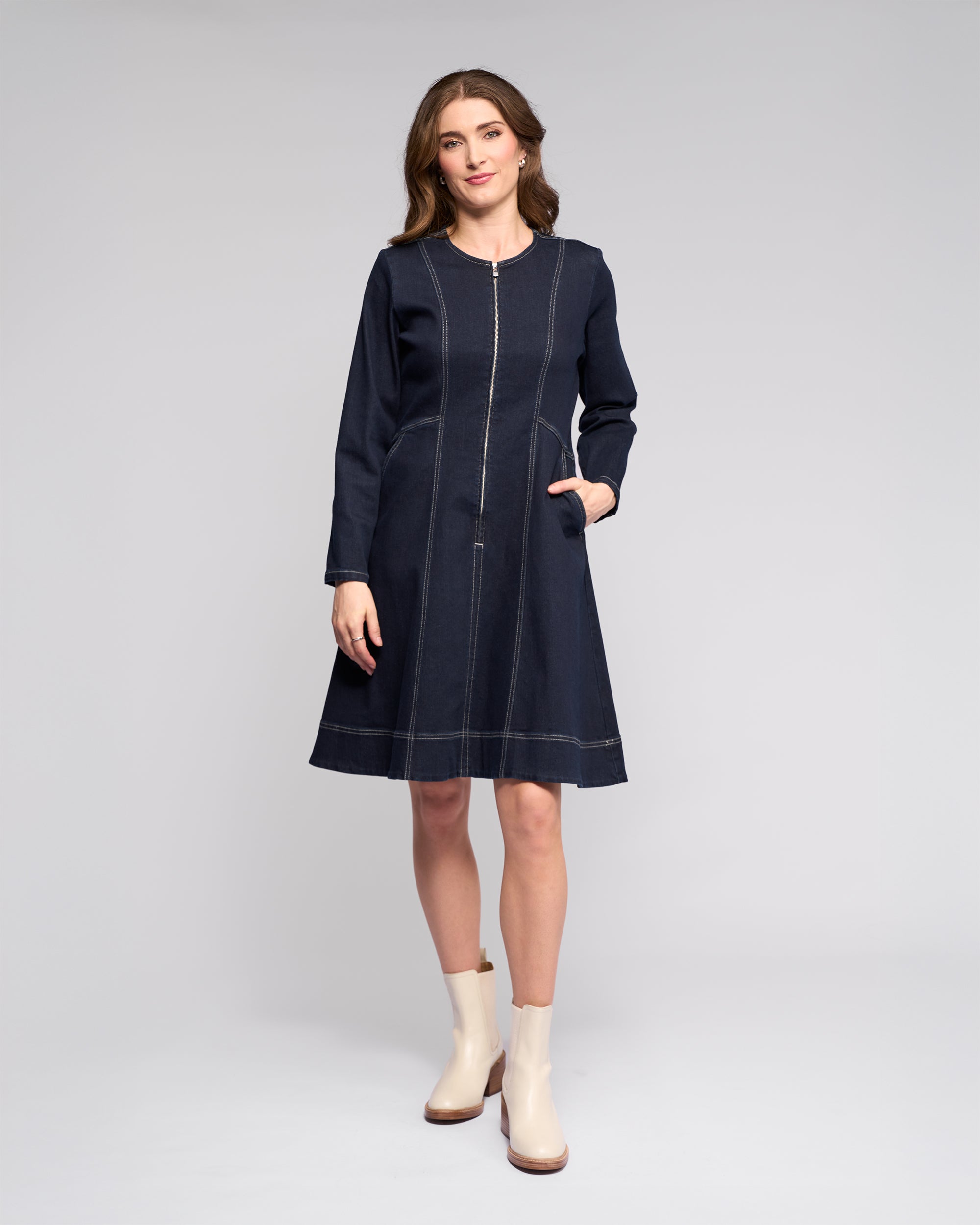 A woman poses in a studio against a light background, wearing the Vassalli Sienna Dress - Ink Denim with long sleeves, visible stitching, side pockets, and light boots, her hand resting in one pocket.