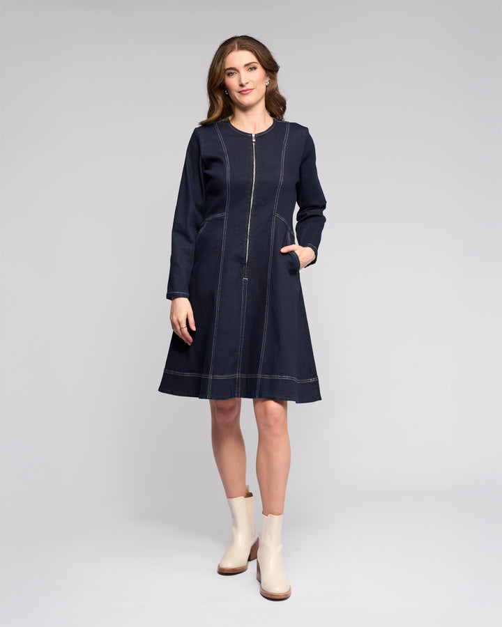 A woman poses in a studio against a light background, wearing the Vassalli Sienna Dress - Ink Denim with long sleeves, visible stitching, side pockets, and light boots, her hand resting in one pocket.